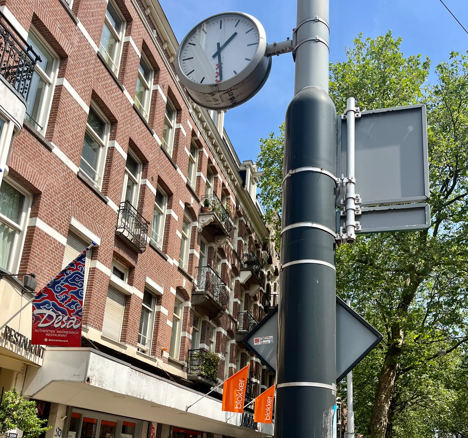 On a bright sunny day, a clock attached to a pole in a shopping street in Amsterdam points at 29 past 1. 
