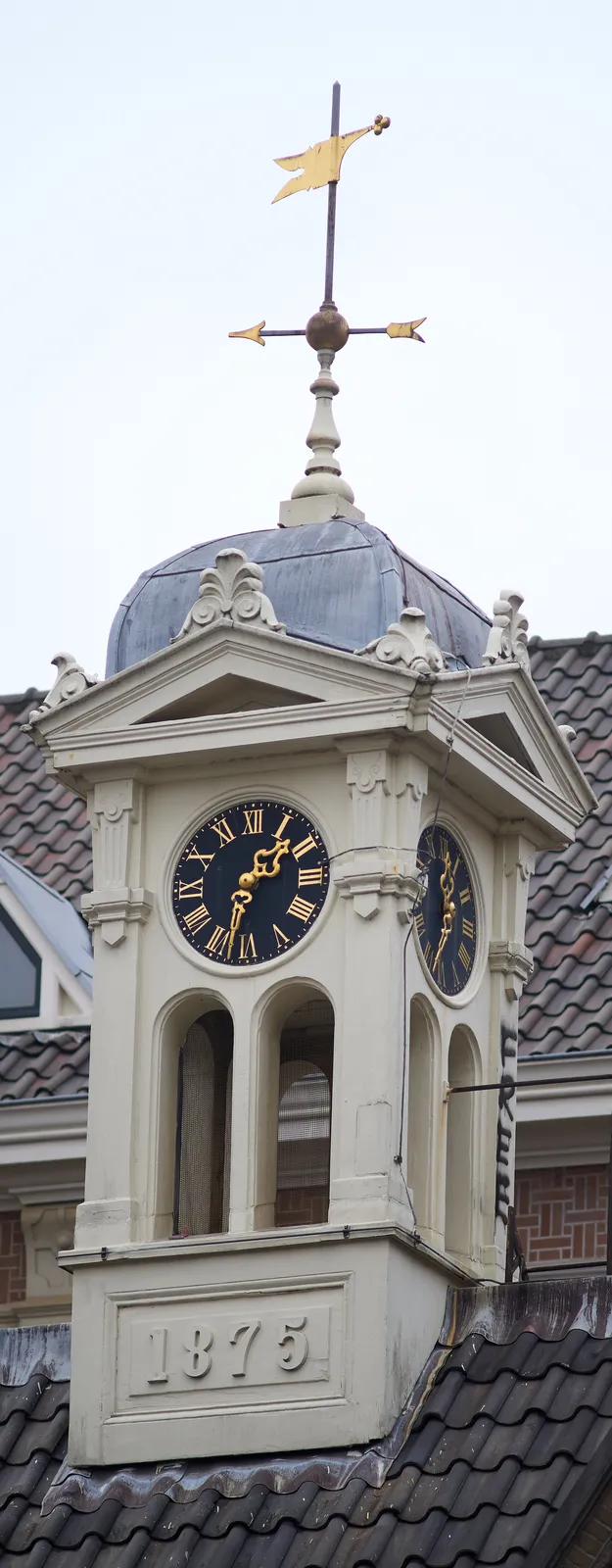 A little tower on top of a roof. The year 1875 is shown at its base. It has some ornaments, and a few small arches. It also has a clock that points at 32 minutes past one.