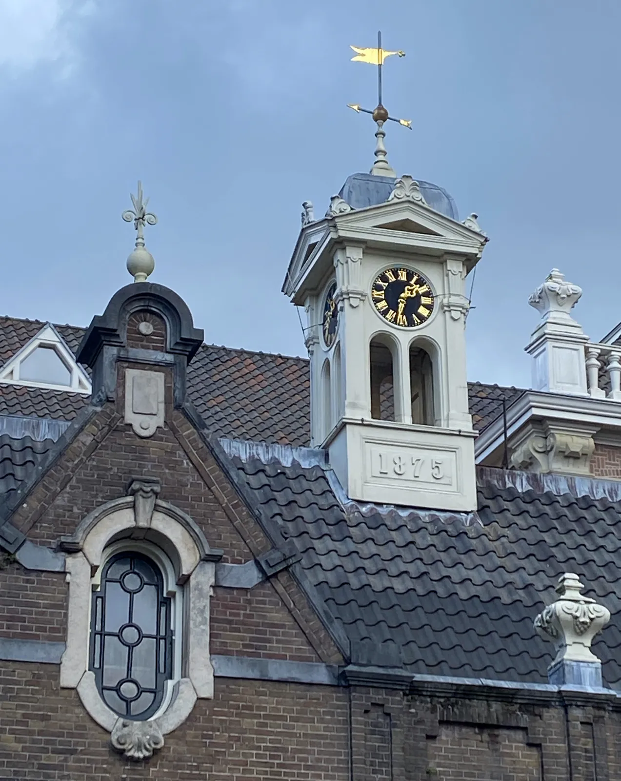 On a roof stands this small, white tower with a wind vane on its top. At the base of the tower it says 1875. The clock on this tower points at 32 minutes past one.