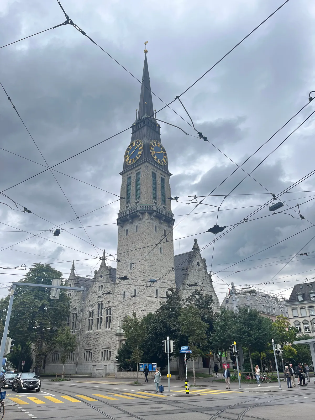 A web of overhead wires for trams in Zürich. Behind the web there’s an enormous church tower with a clock on it. It points at 19 minutes to two.