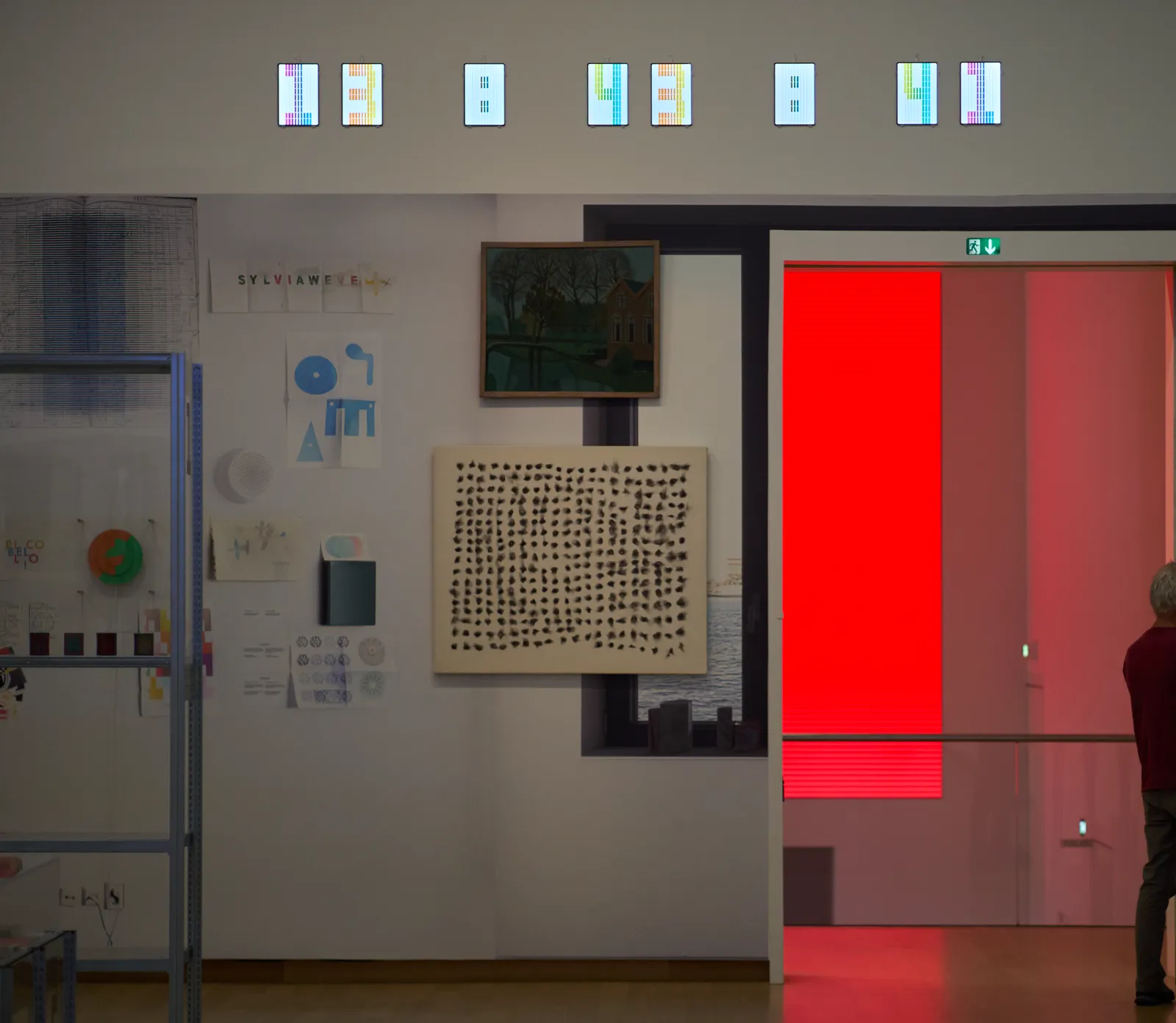 Very many works of art hanging on a wall with a large door in it, leading to another space with red light. Above this door are eight small displays, all displaying a number or a colon. Together they display the time as 13:43:41