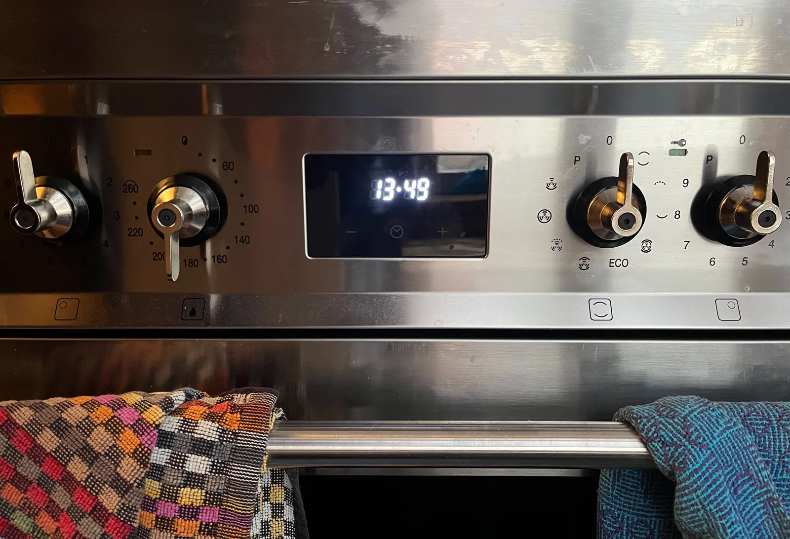 The four mechanical knobs of a metal, electrical oven. There are towels hanging over the door rail. There’s a digital clock between the knobs which says that it is 13:49