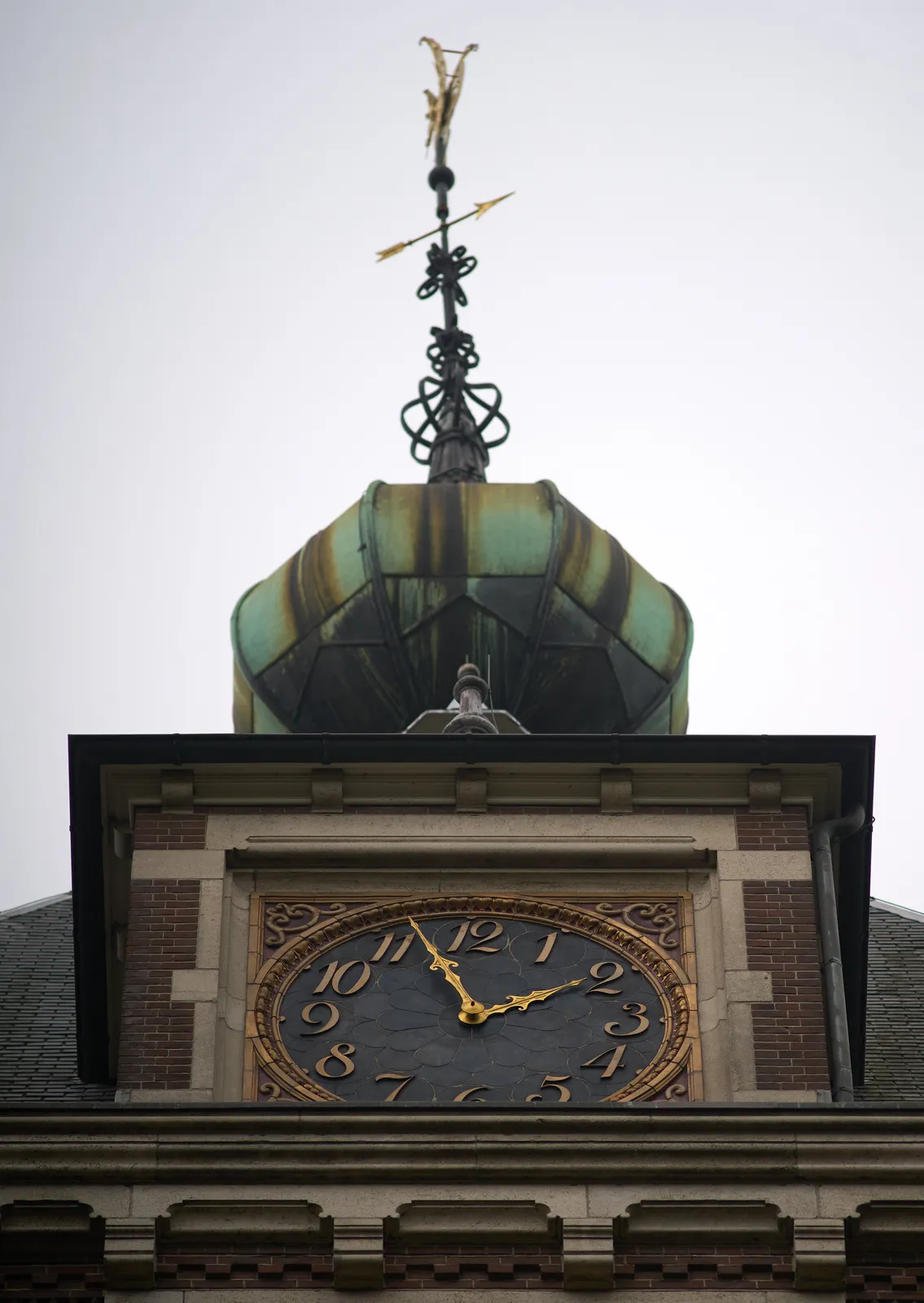 Looking up at a tower. The tip is made from copper. Below this tip there’s a large clock bricked into the wall. The clock is nicely ornamented, and it had golden hands that point at 9 minutes to two.