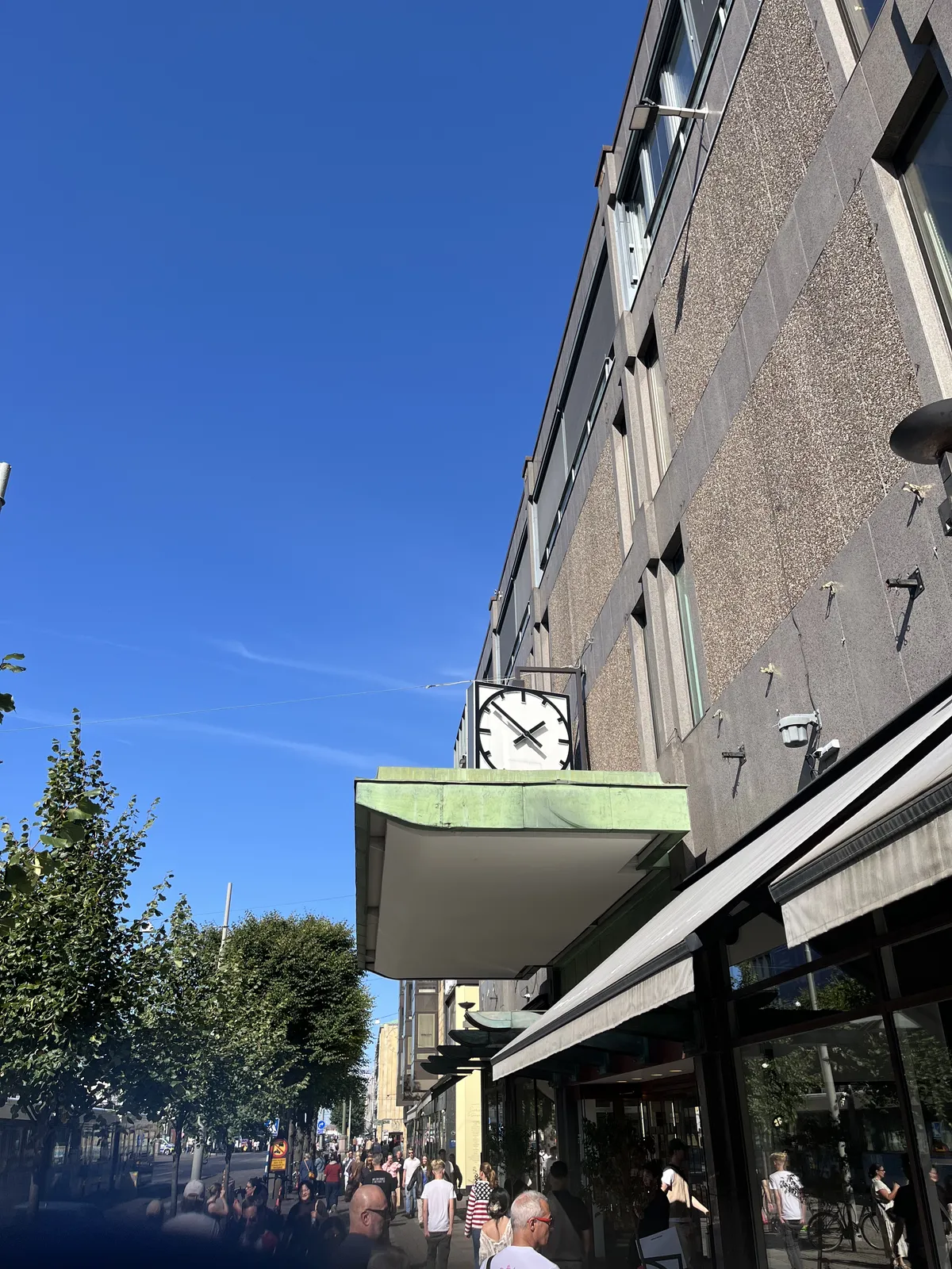 Attached to a concrete building in a rather busy shopping street is a large modernist clock that points at 9 minutes to two.