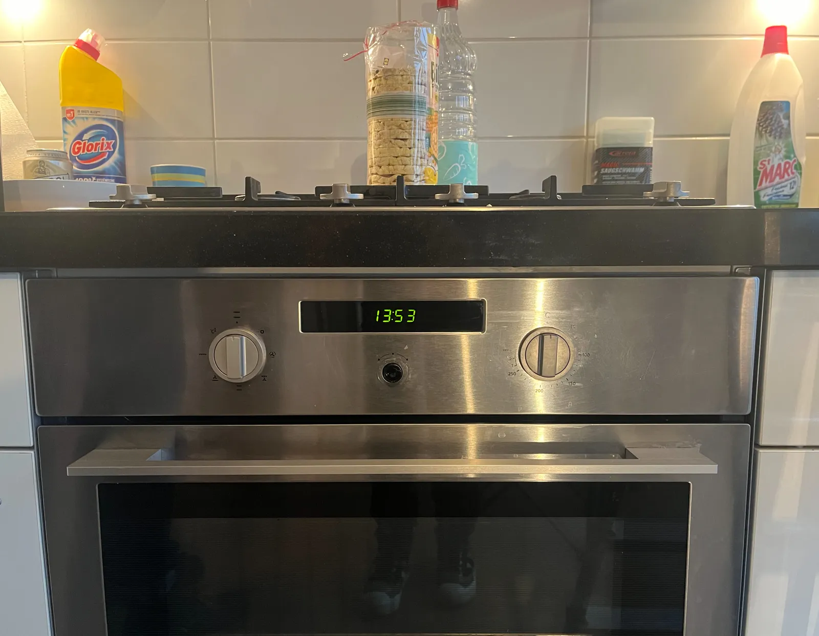People are cleaning in this kitchen. Lots of different bottles of detergent. There’s an oven with a digital clock on it. According to this clock it is 13:53