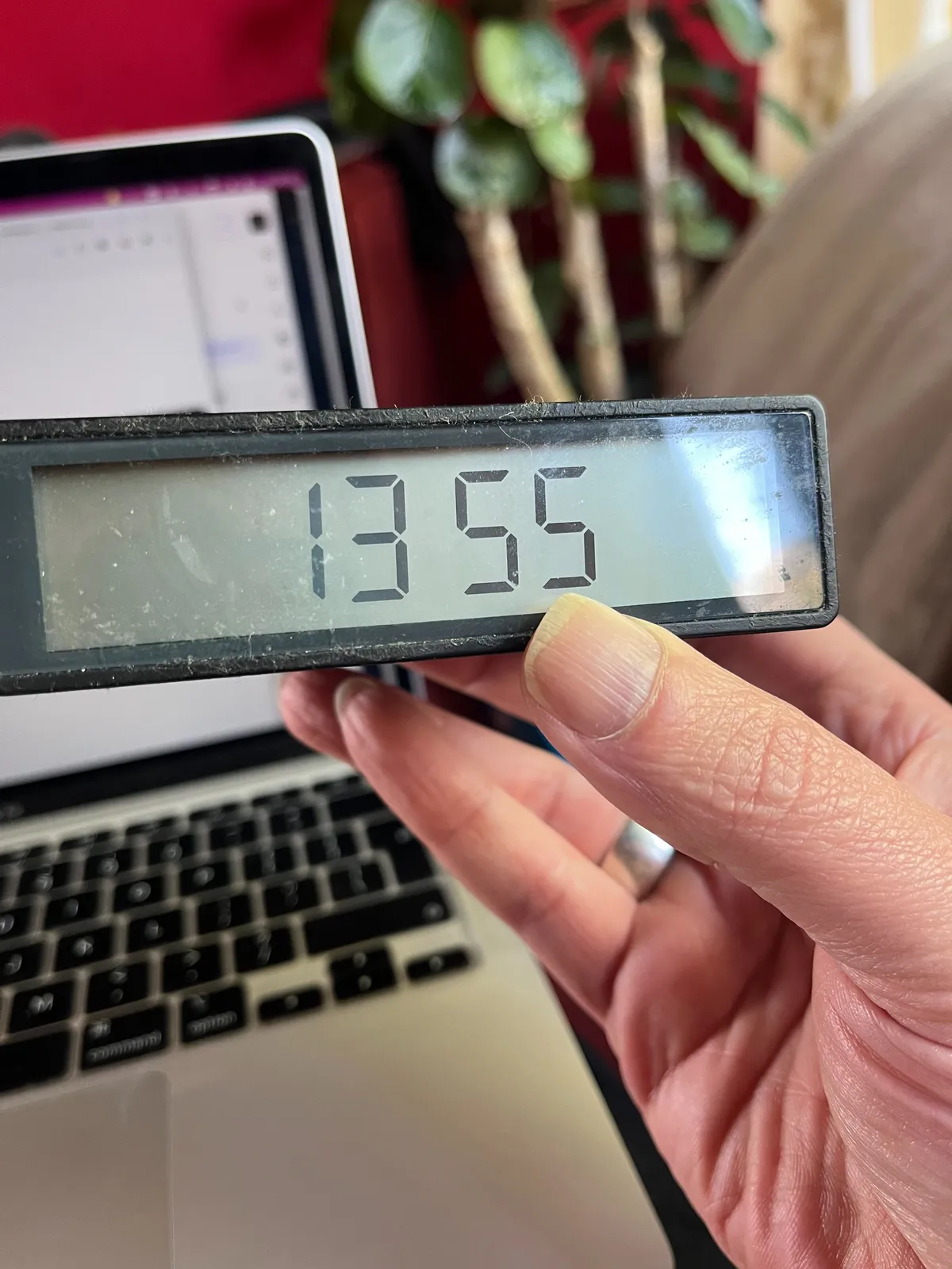 Someone is holding a digital clock in front of a laptop computer. In the background there’s also a plant. The clock shows the time as 13:55