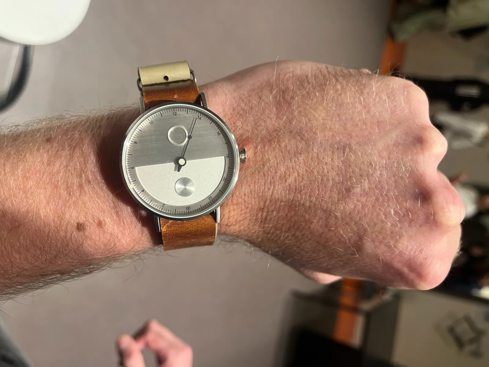 A minimalist wrist watch. It has one single hand, the hour hand, which turns just once every day. The bottom of the face is made of a lighter coloured material than the top. When you look closely at the tip of the hand, you can see that it points at approximately 3 minutes to 2.