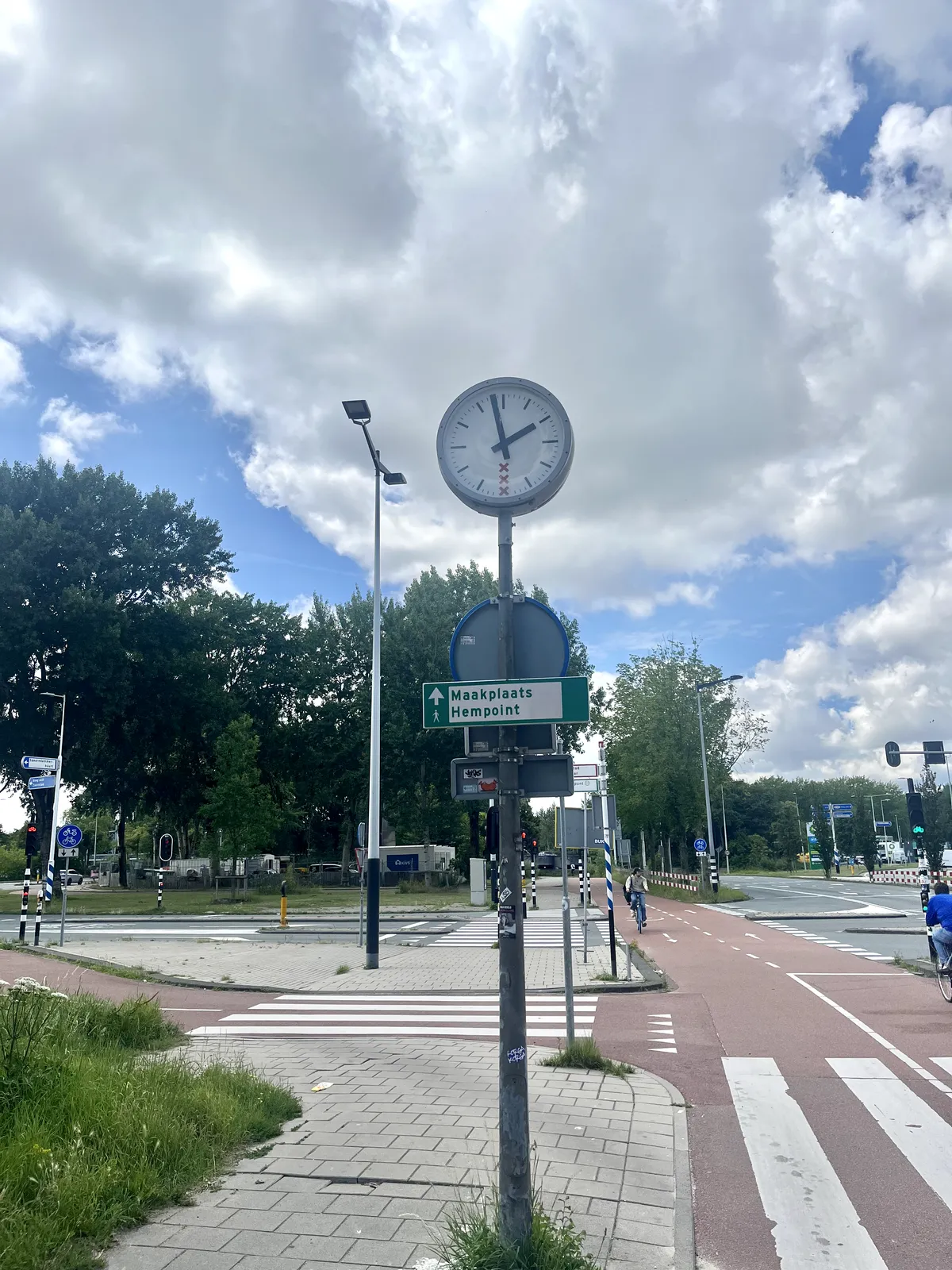 A clock on a point next to a bike lane. There’s a sign below the clock that points in the direction of Makerspace Hempoint. The clock points at 2 minutes to two.