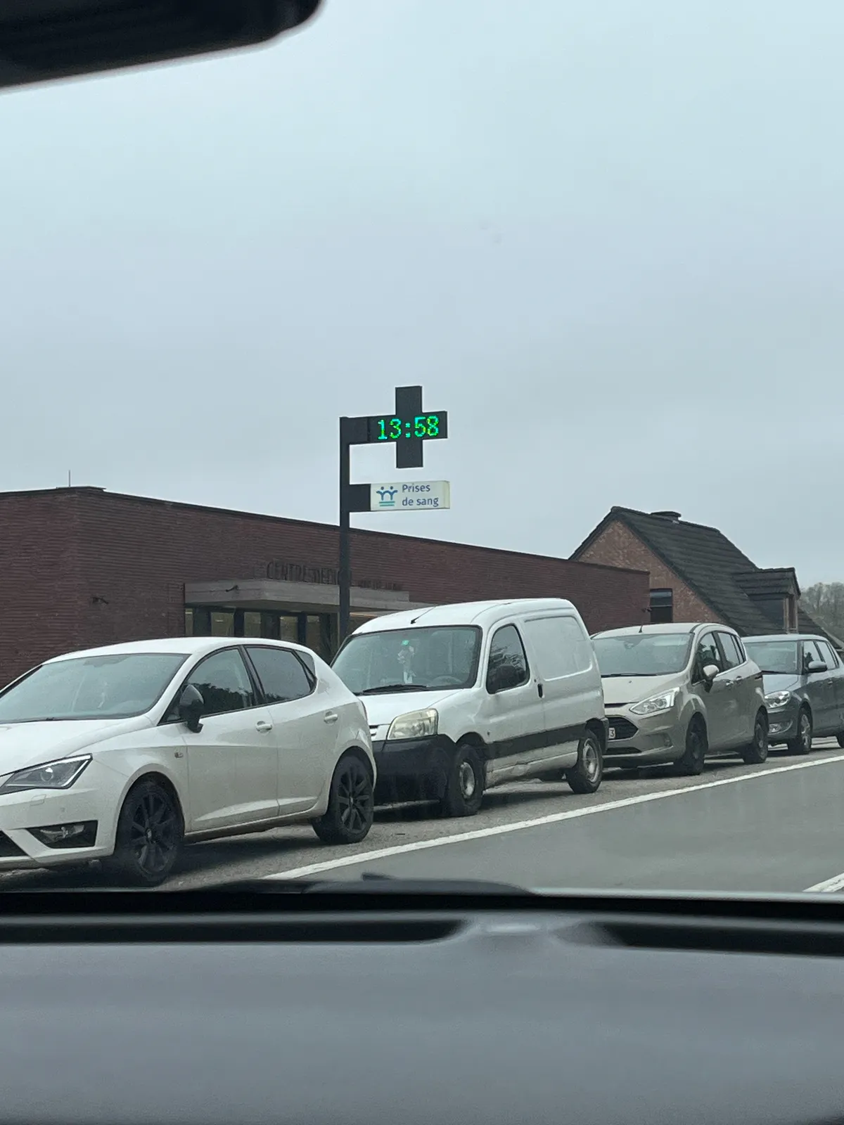 Four cars are parked in front or a square brick building. There’s a pole behind these cars with a cross shaped display on it. The time is shown on this display as 13:58.