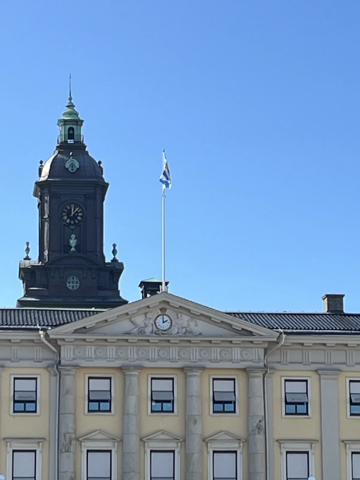 Behind a yellow, neo-classical building there’s a dark chrurch tower with a clock on it which points at 6 minutes past twelve. On the neo-classical building there’s a clock that points at two o’clock.