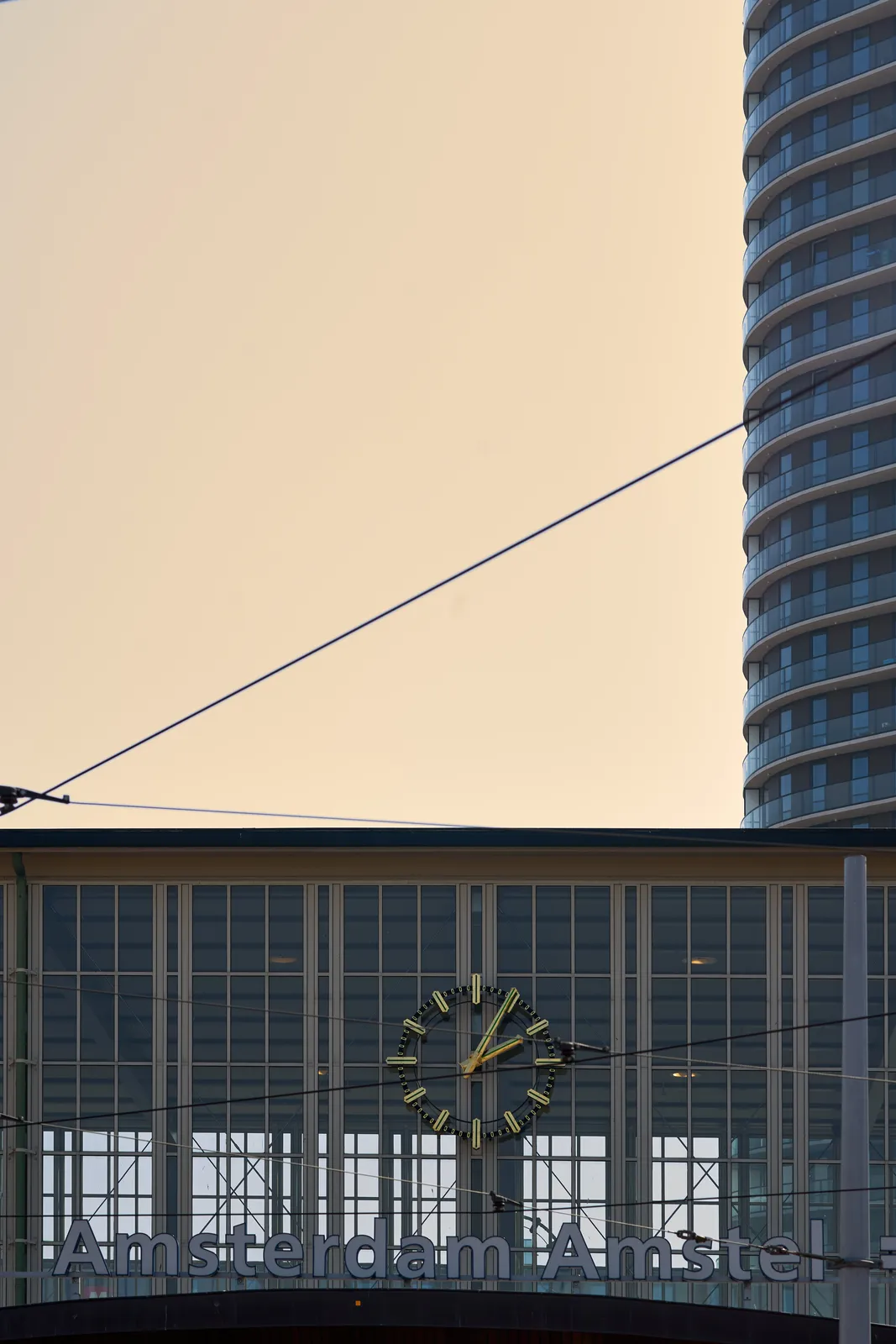 In the background a skyskraper can be seen. In the foreground is the Amstel Station in Amsterdam. The large clock above the entrance points at almost 5 minutes past five.