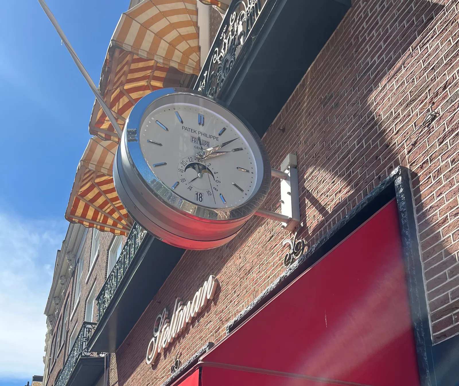 A large, very shiny clock is attached to the facade of a building. It says Patek Philippe on its face. Apart from the time, it also shows the date, Friday 18 April, and it shows if it’s day or night, which can also be seen by looking at the bright blue sky. The clock says it is 7 minutes past 2