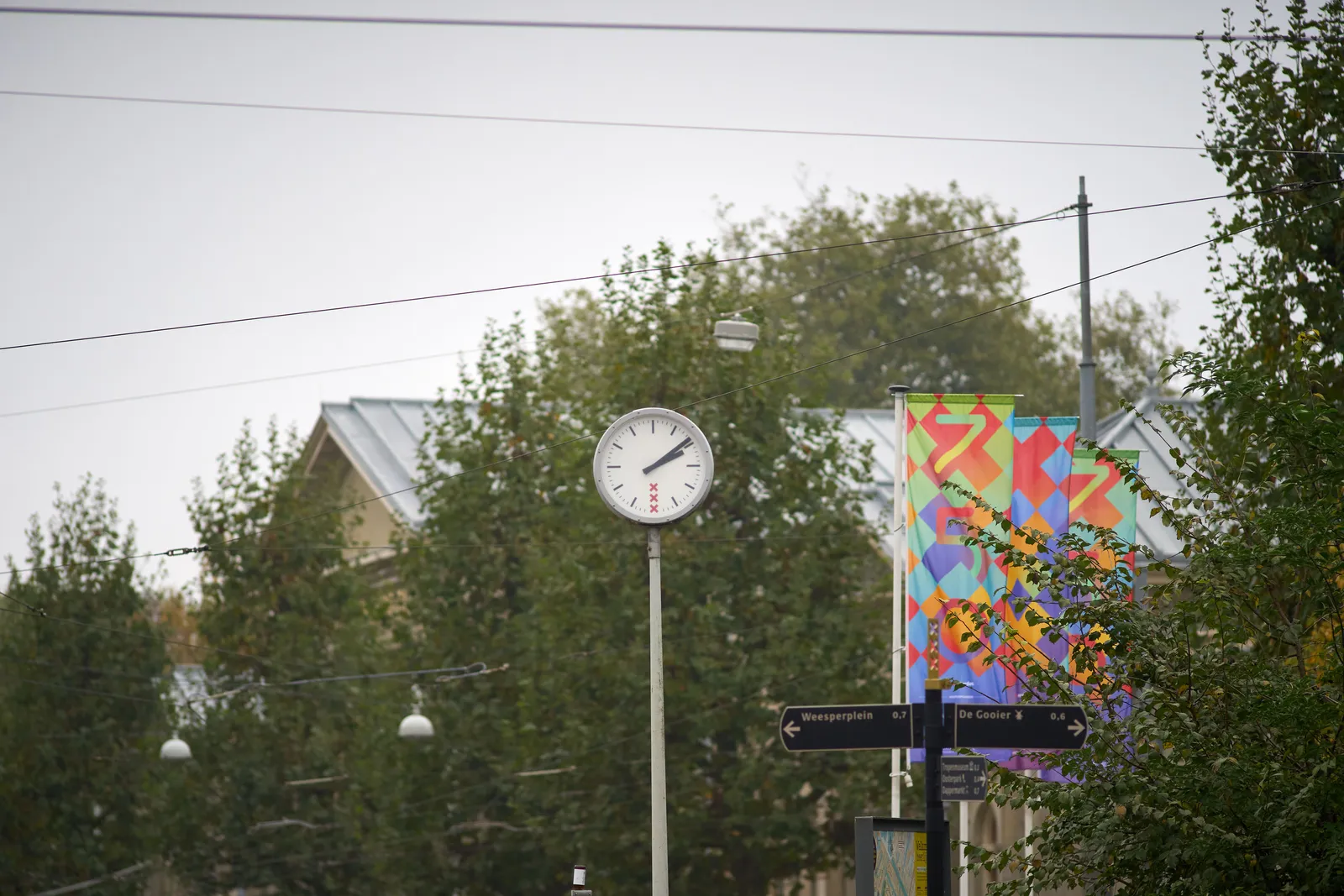 A clock high up on a pole on front of some trees. There are also some very colourful flags in this picture. The clock points at 9 past 2.