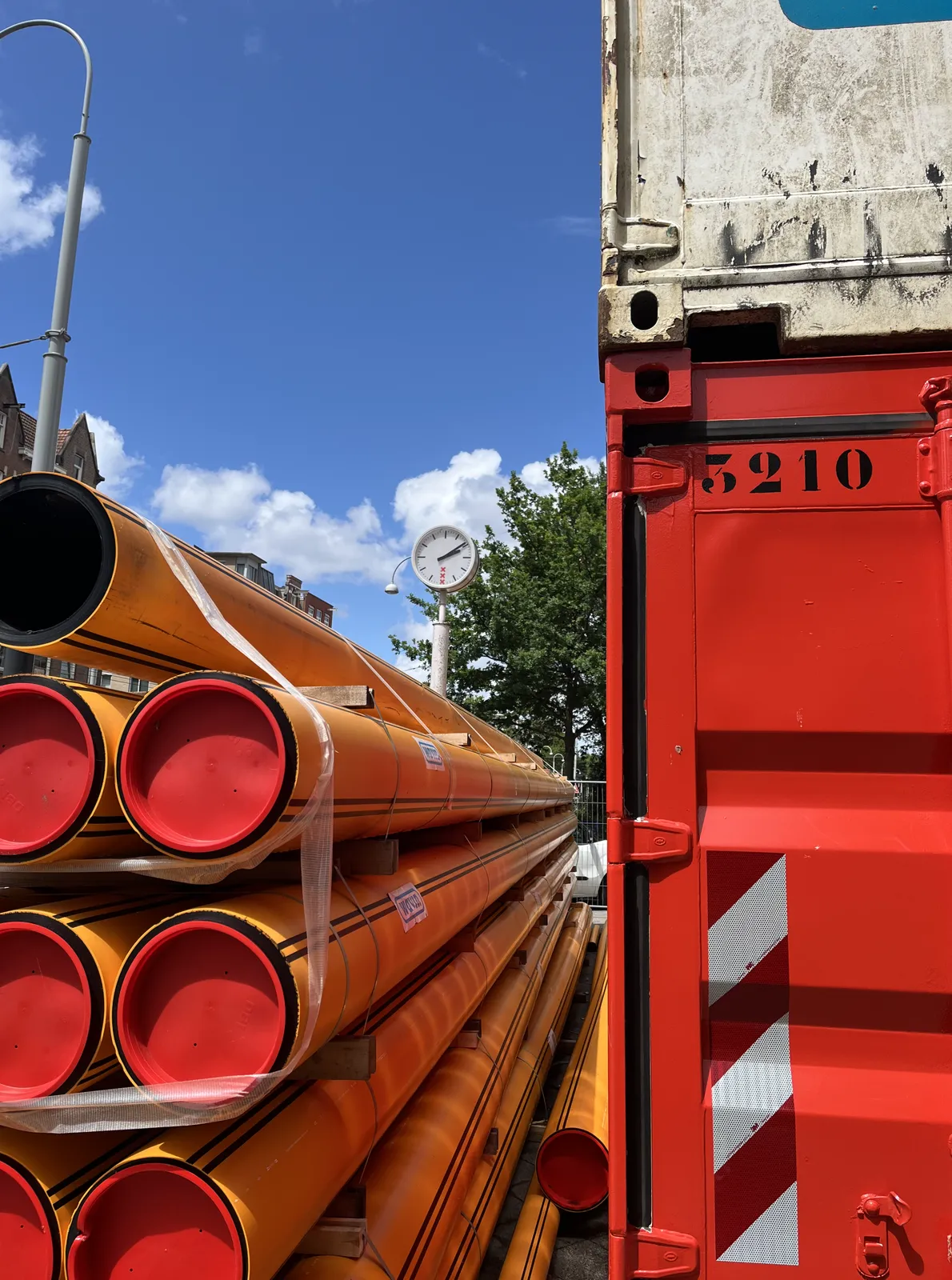 Looking at a bright red container, with bright orange pipes next to it, against a bright blue sky. Behind the pipes there’s a clock on a pole that points at 9 minutes past two.