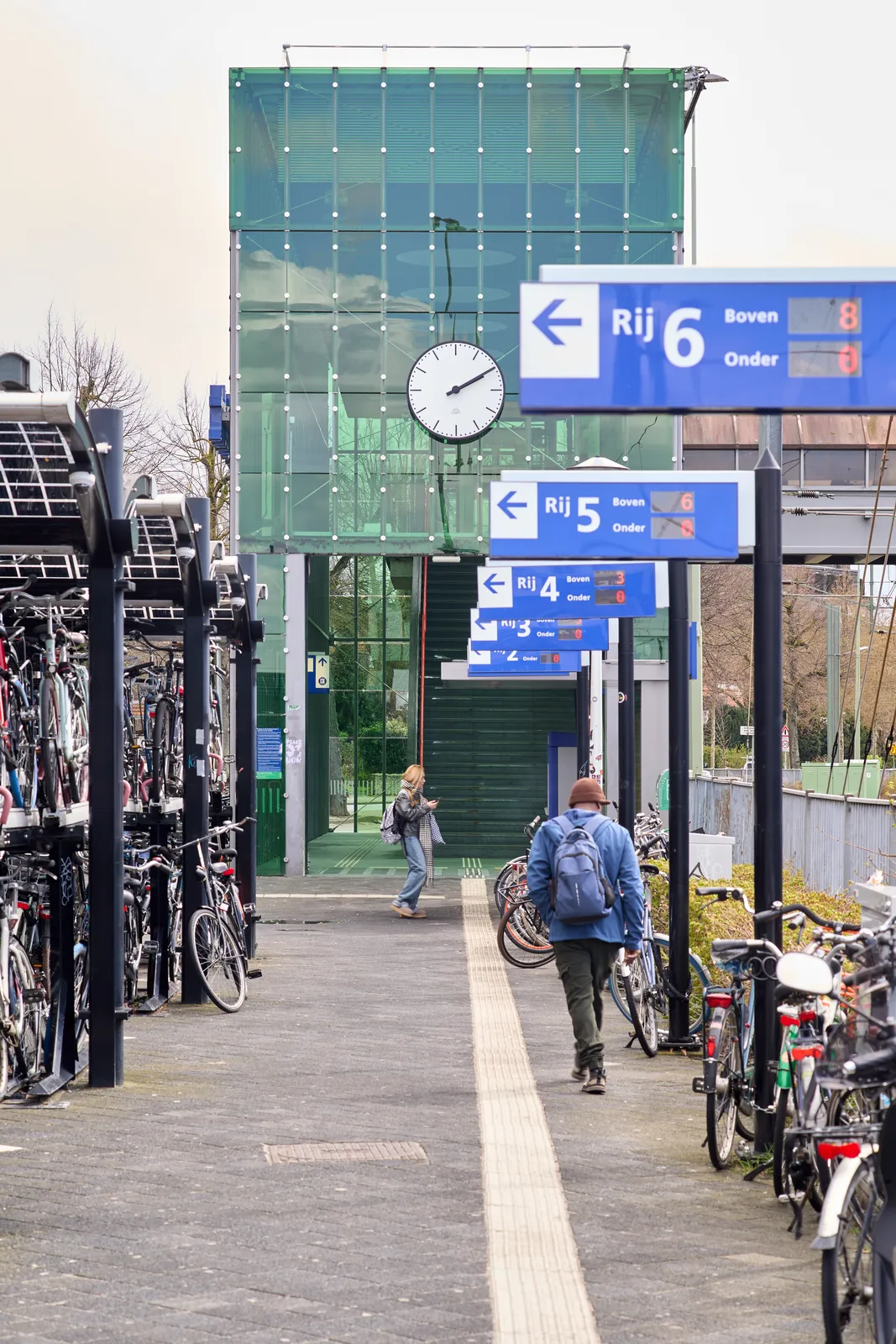 An array of signs that point at row number 2 to six. Behind this array there’s a stairway in a towerlike building made from glass. On its facade there’s a clock that points at 10 past two.