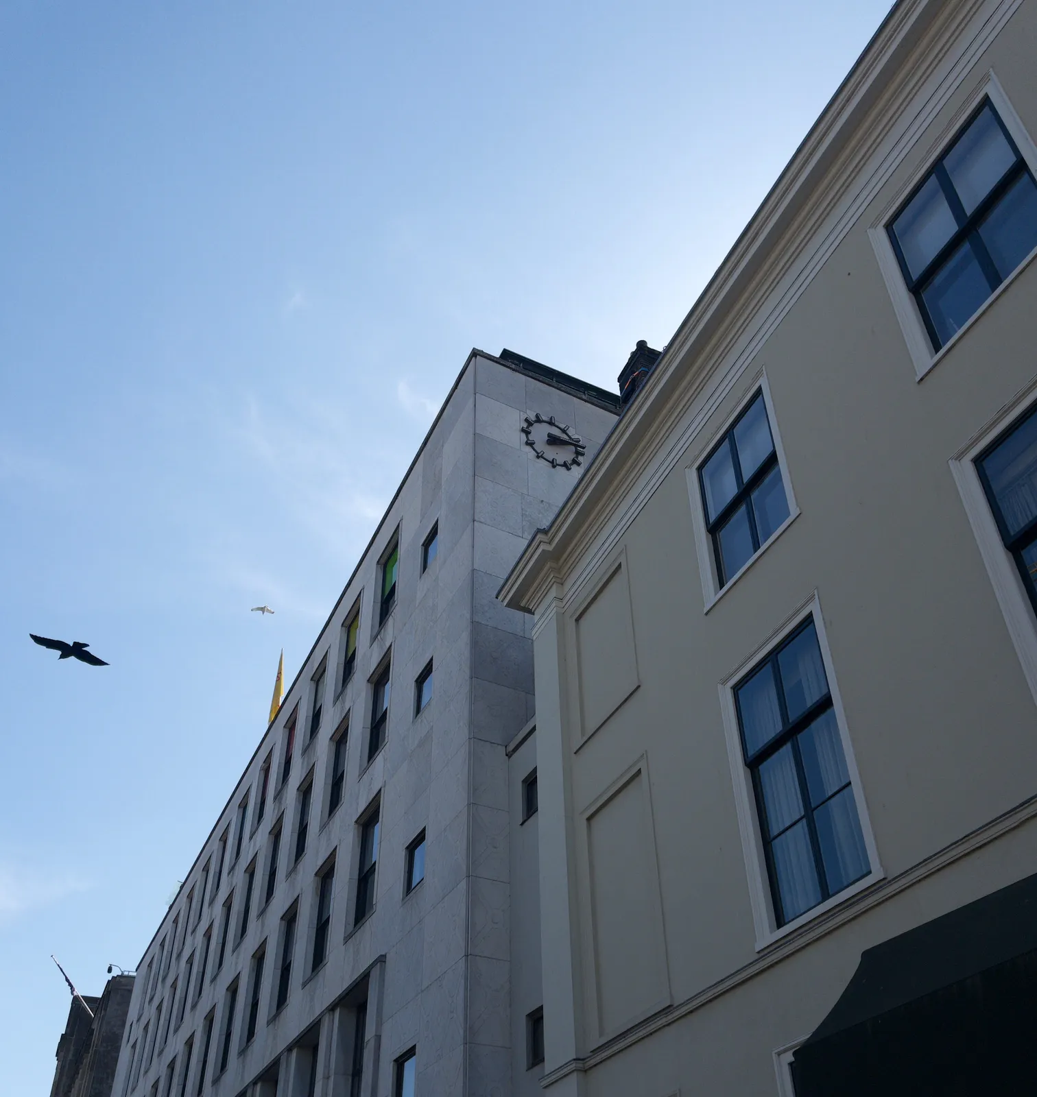 A bird is flying past two buildings. A more classical building in the foreground, and behind it a modernist building. There’s a clock on this more modern building, which point at 13 minutes past 2