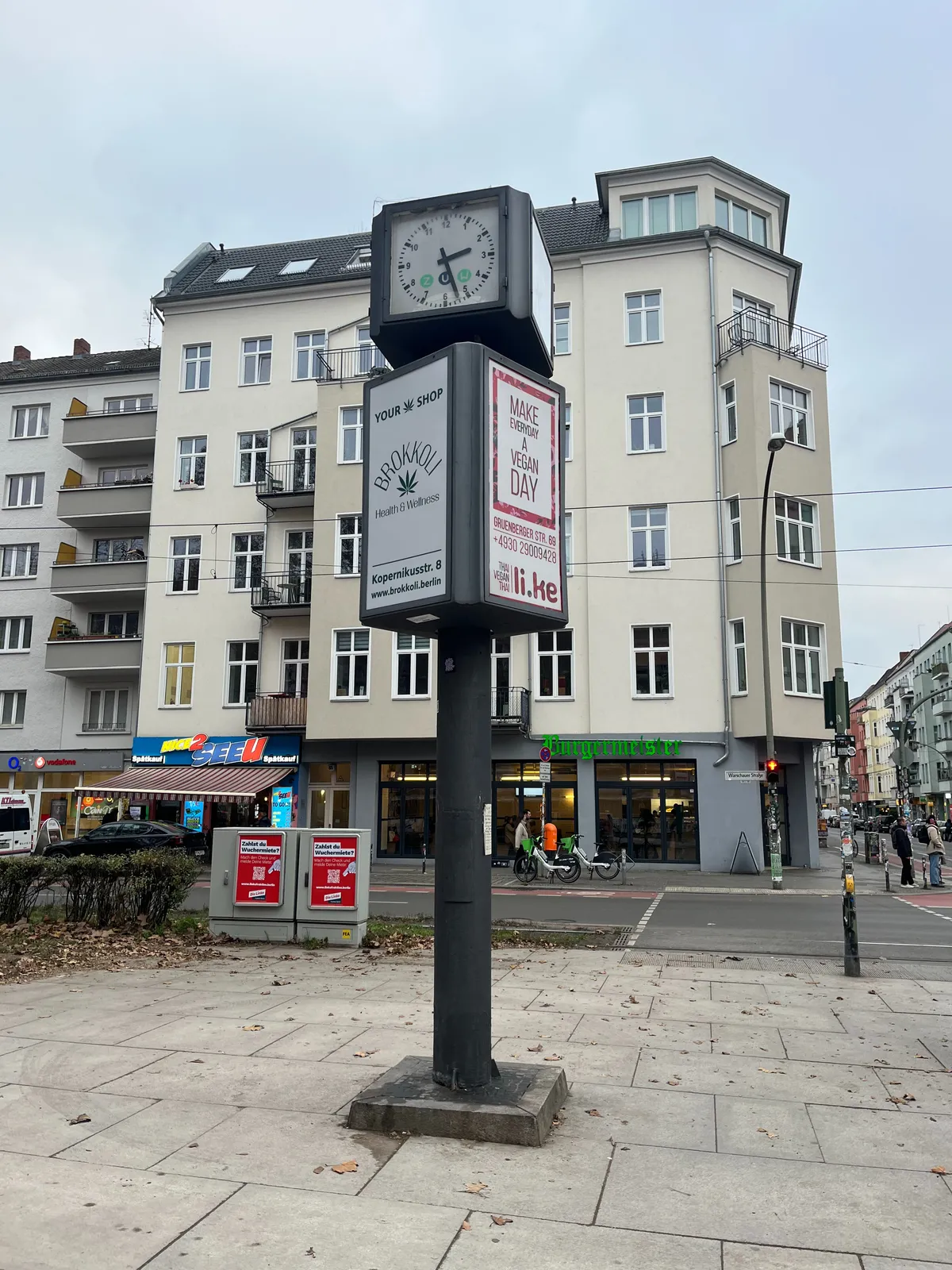 A rather high appartment building in the background. In the foreground there’s a high pole with advertisements on it. The one on the left is for Brokolli, which seems to be a marihuana shop. The one on the right tells us to make every day a vegan day. On top of this advertisement pole there’s a clock that points at 22 minutes past 2.