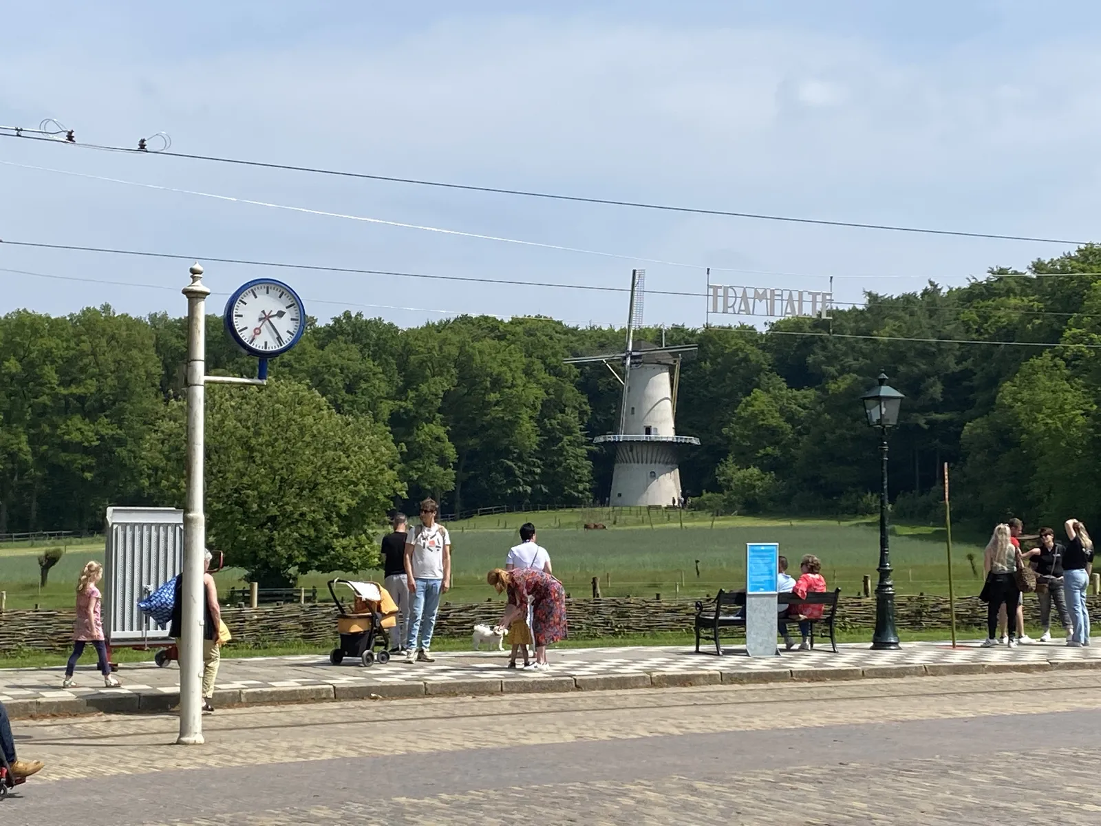 In the background there’s a forest and a windmill. People are waiting at a tram stop. There’s a clock on a pole that points at 24 past 2.