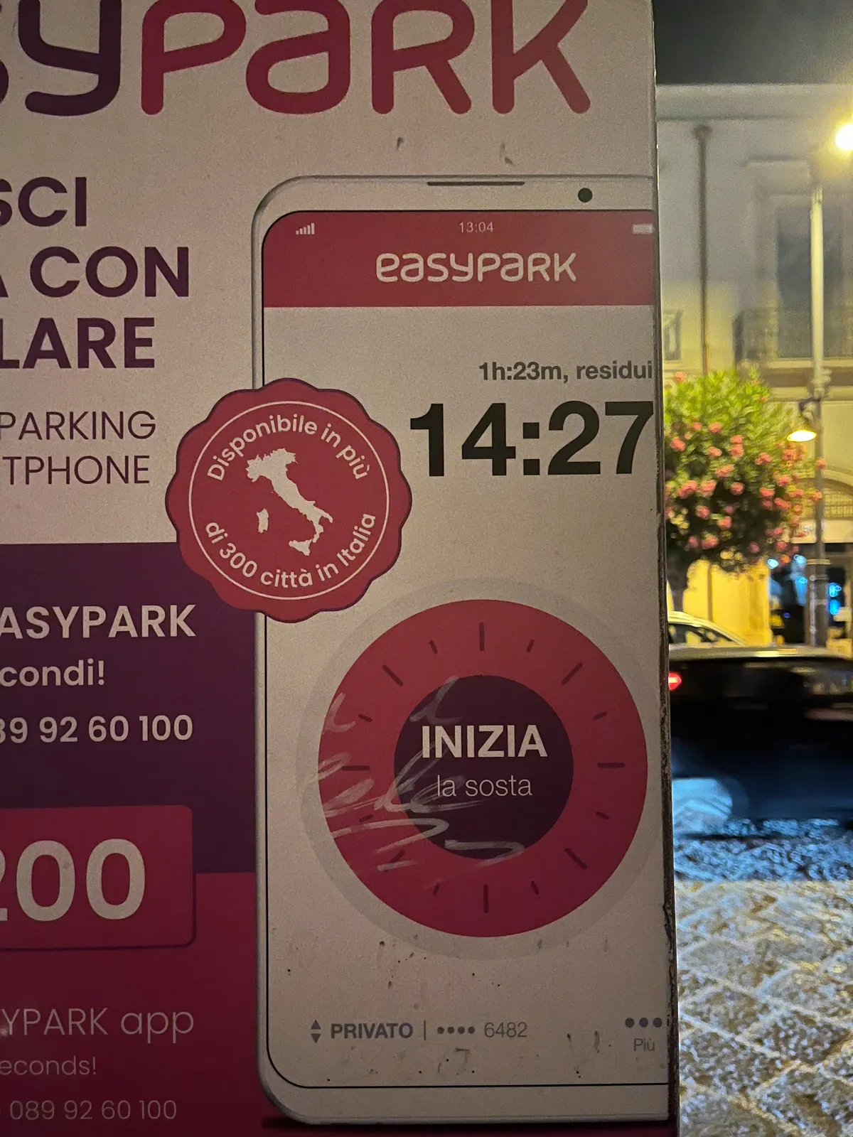 An advertisiment for a parking app on a street, at night. There’s an abstract idea of a phone on this advertisement, with the time printed on it as 14:27.