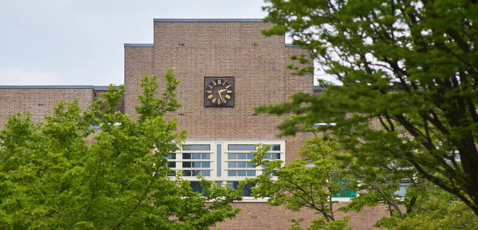 A large clock on something that looks like an old brick school building, surrounded by trees. The clock points at 27 past 2. 