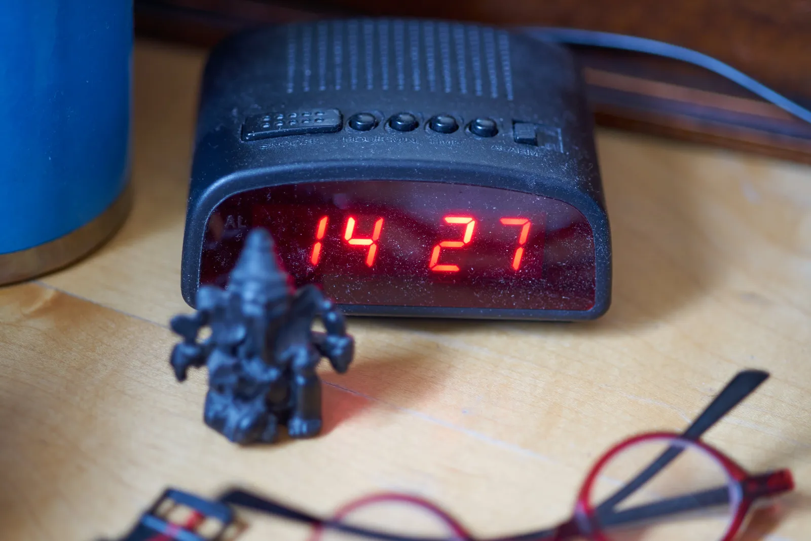 A digital alarm clock, surrounded by a little statue, some books, and red reading glasses. On the clock the time is 14:27