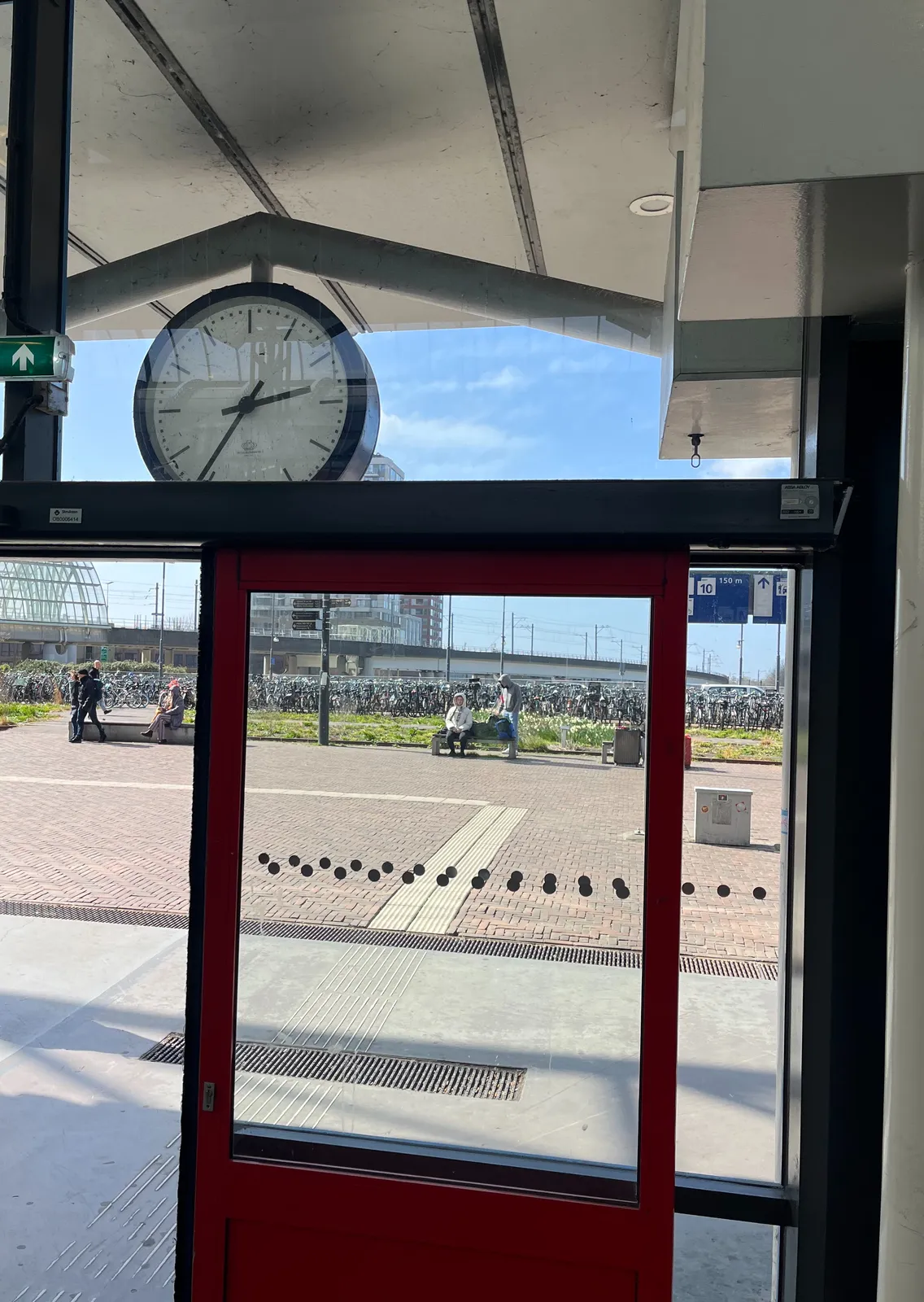 Looking out through the glass facade of a Dutch train station, at a square on a sunny day. Behind the door there’s a station slock which points at 36 minutes past 2.