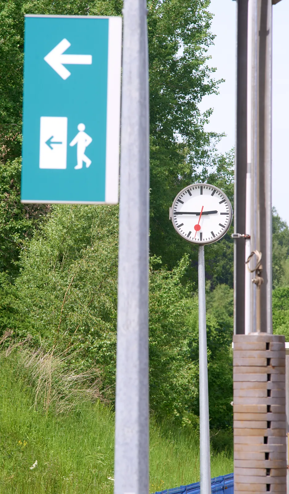 Outside, surrounded by green nature, there are a sign that points at an exit, a heavy weight, and a station clock which points at a quarter to three.