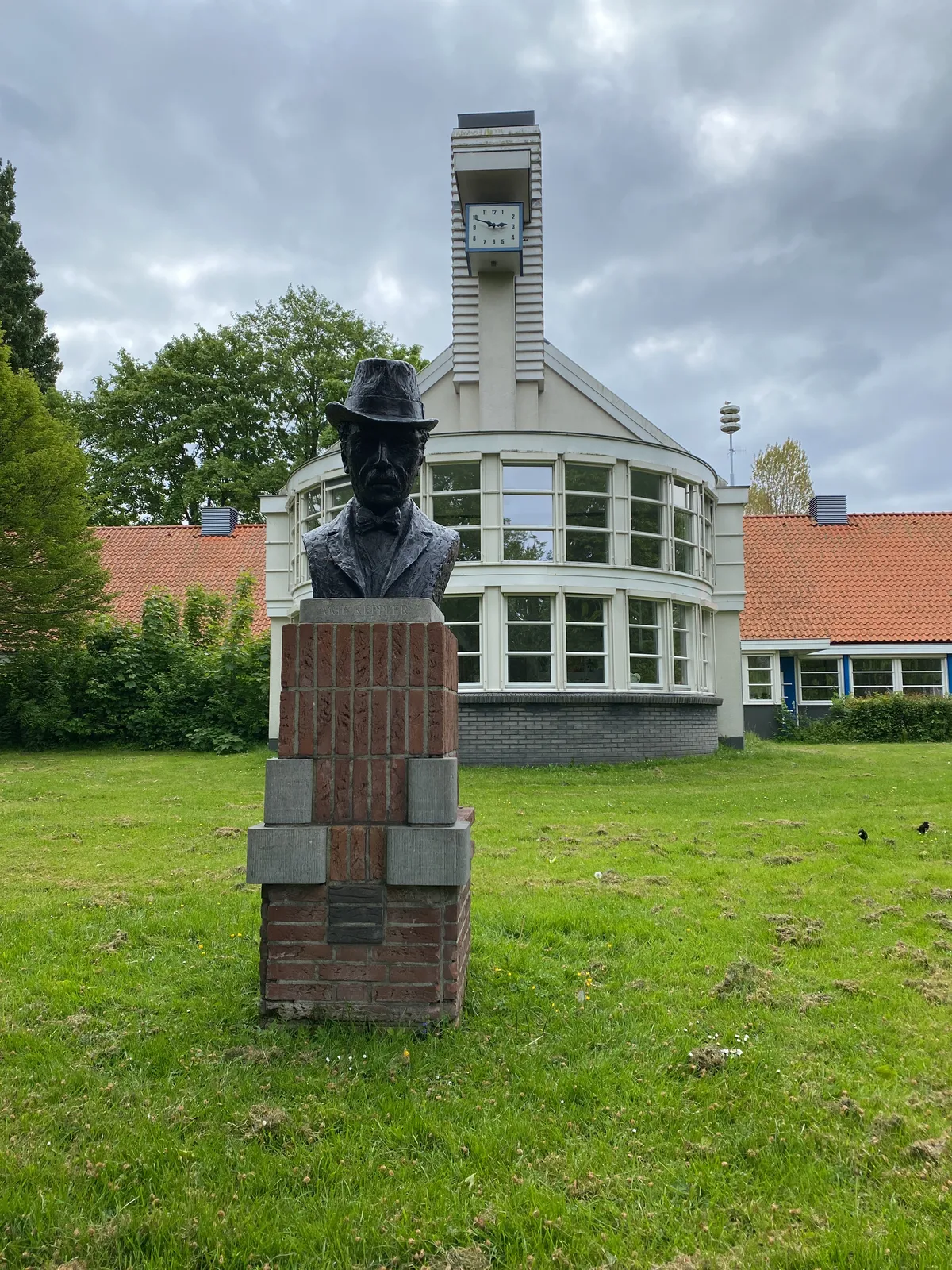 A statue of a man with a hat. Behind the statue there’s a modernist building with a clock on it. It points at 11 to 3.