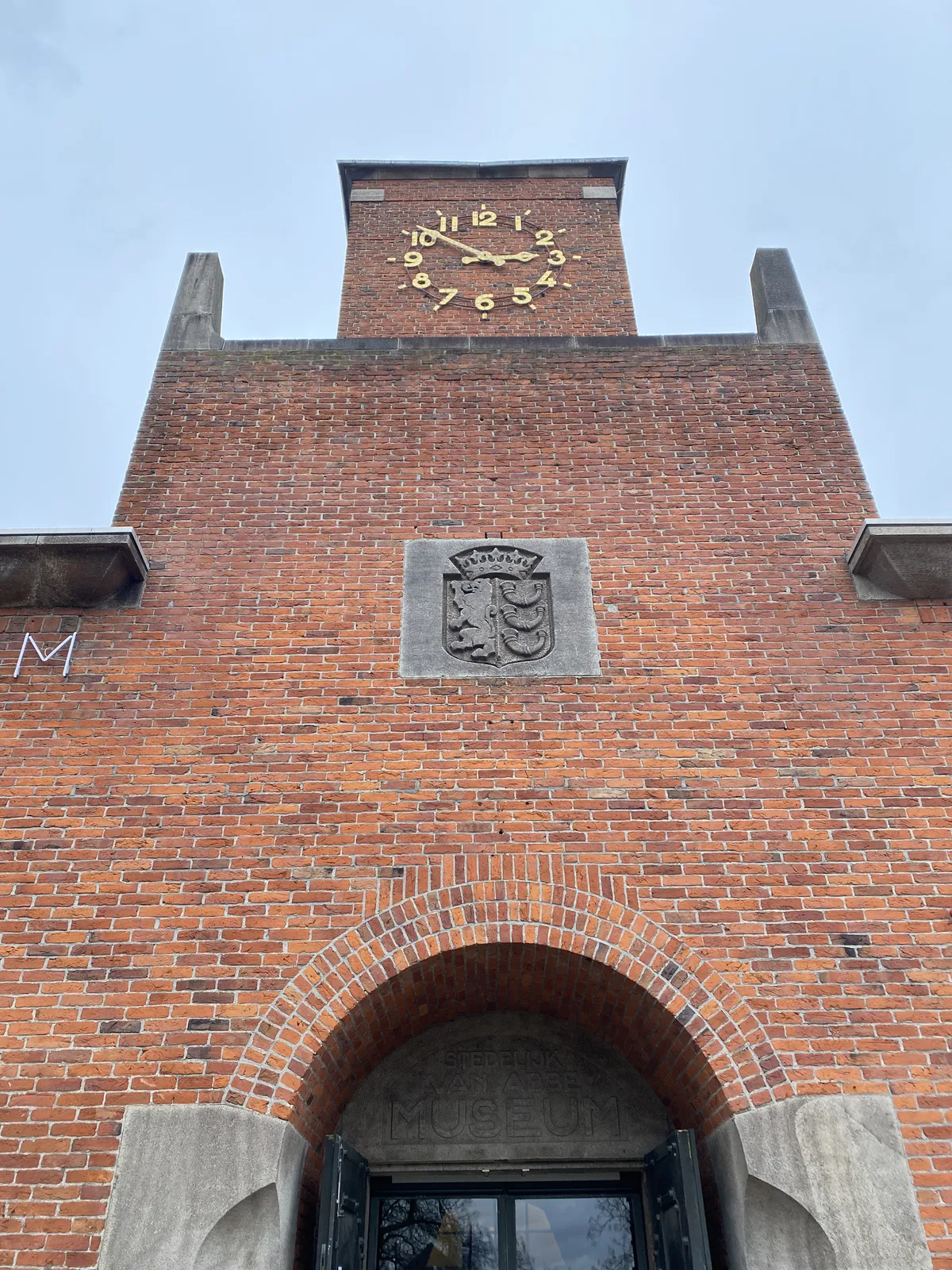 A symmetrical picture of the entrance of the Van Abbemuseum, it says so in art deco letters on the facade. At the top of the building there’s a clock. It points at 9 minutes to 3.