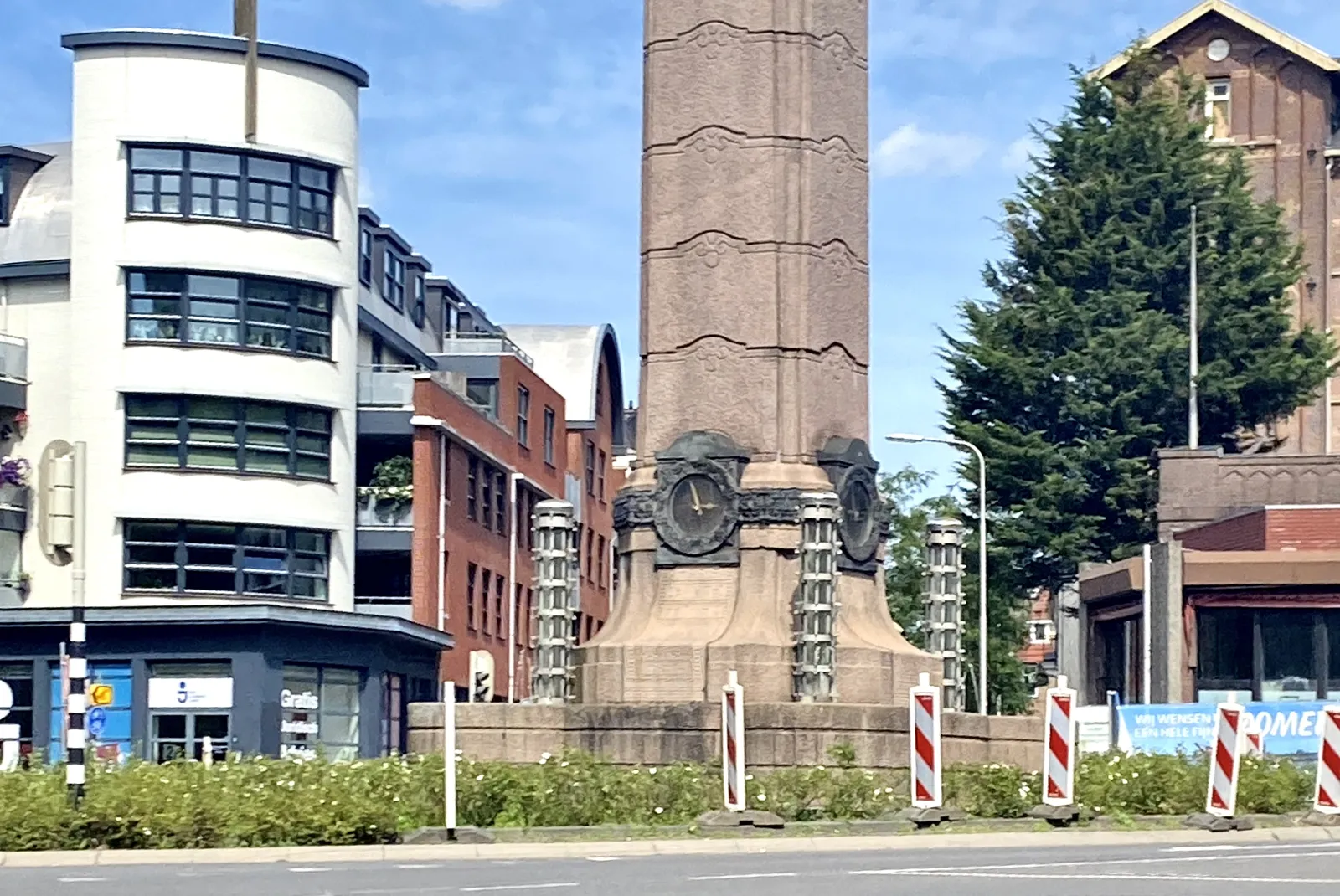 A large column with a clock at its base, in front of some houses. The clock points at 4 minutes to 3.