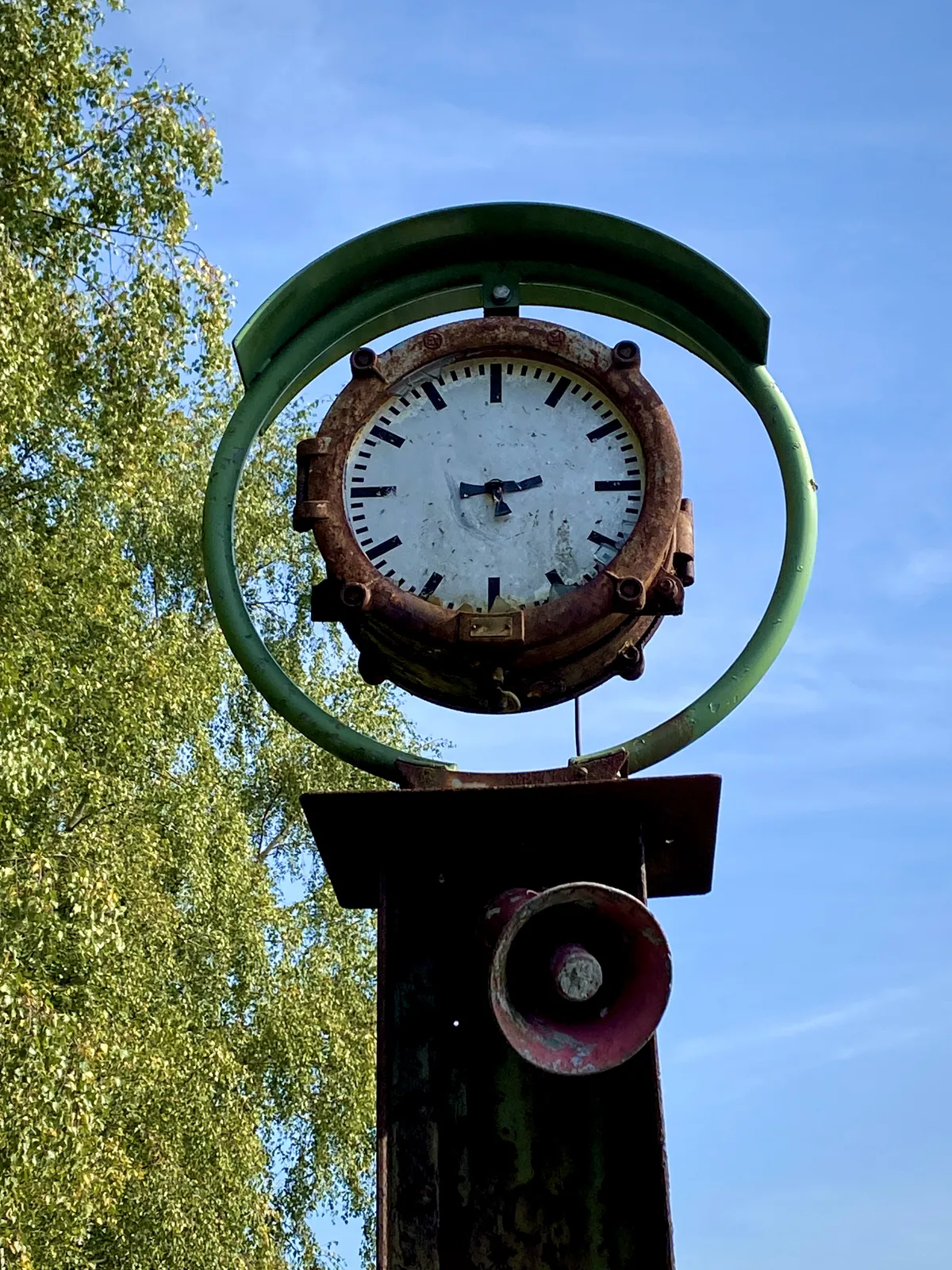 A clock in a green round frame, mounted on a simple piece of construction metal. An alarm horn hangs below it. The hands of the clock are broken, so it’s hard to tell what time it is exactly. But if you look closely it seems to be pointing at 3 minutes to 3. 