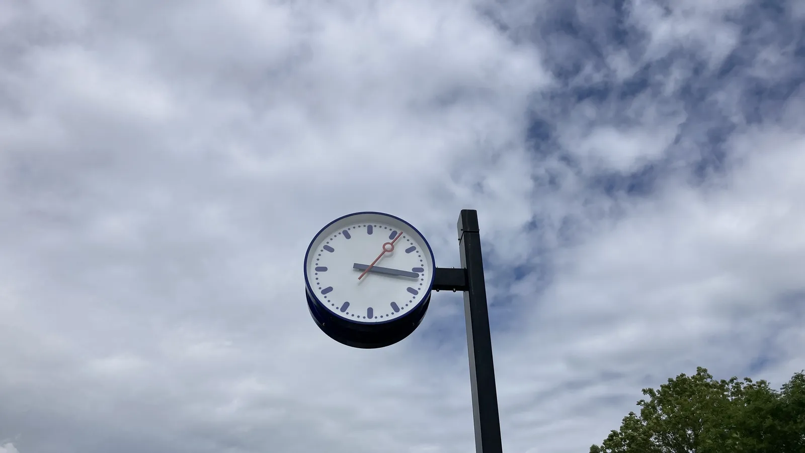 A cloudy summer day. A tree in the right bottom corner. In the center there’s a clock on a pole. It points at 16 minutes past 3.