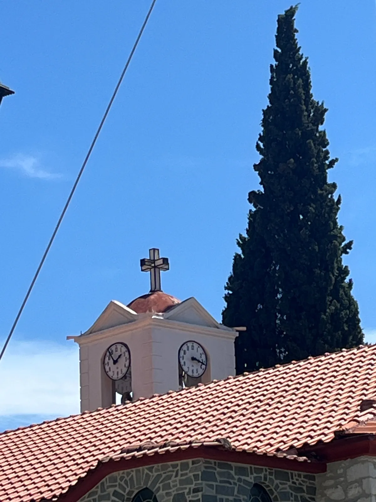 Behind a small bizantine shaped church there’s a tall cypress tree. The church has a little tower with two clocks on it. One of the clock points at 8 minutes to 2, the other points at 17 minutes past 3