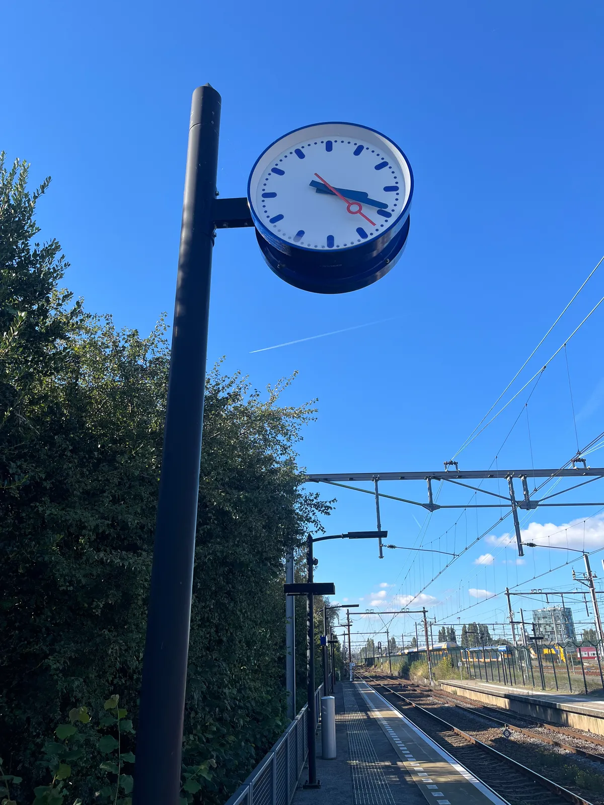 Looking past the train tracks. On the platform there’s a clock on a pole which points at 19 minutes past 3.