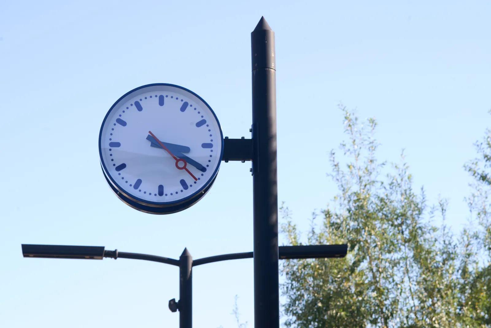 A clock attached to a pole. There are also some streets lights and a tree in the picture. The clock points at 20 minutes past 3.