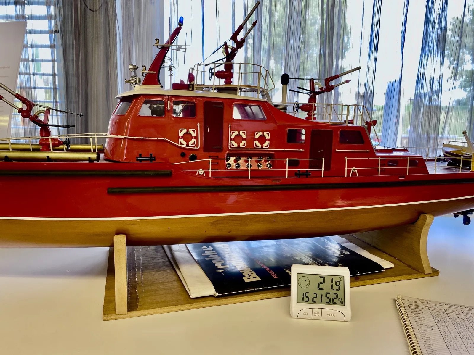 A model of a red boat. In front of this model there’s a digital clock, that shows the humidity in the room, which is at 52%, it shows the temperature, which is 21.9 degrees, it shows a smiley face, and it displays the time as 15:21.
