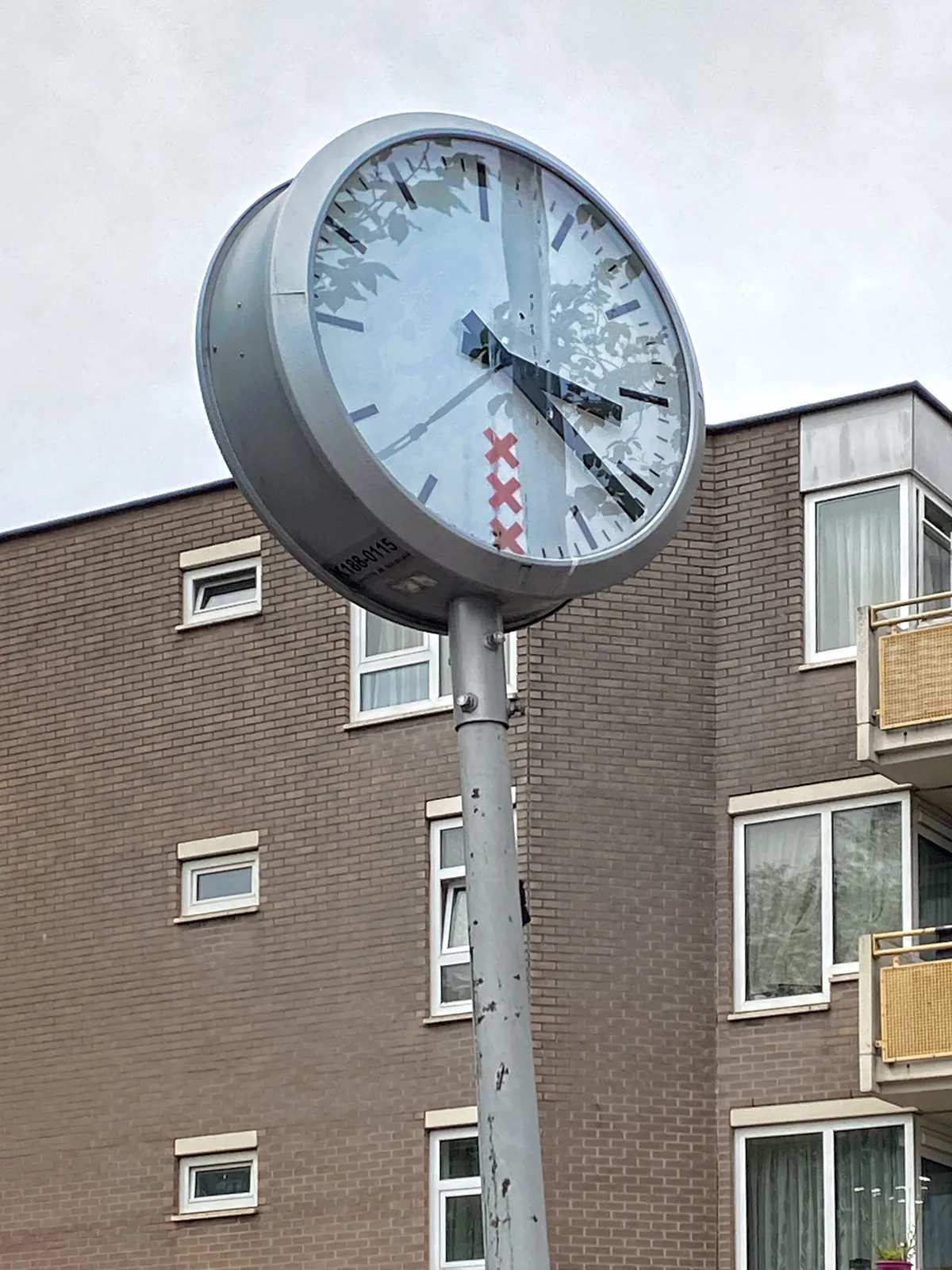 A modern, brick apartment building in the background. In the foreground a clock with the logo of the municipality of Amsterdam on its face. It points at 21 minutes past 3.