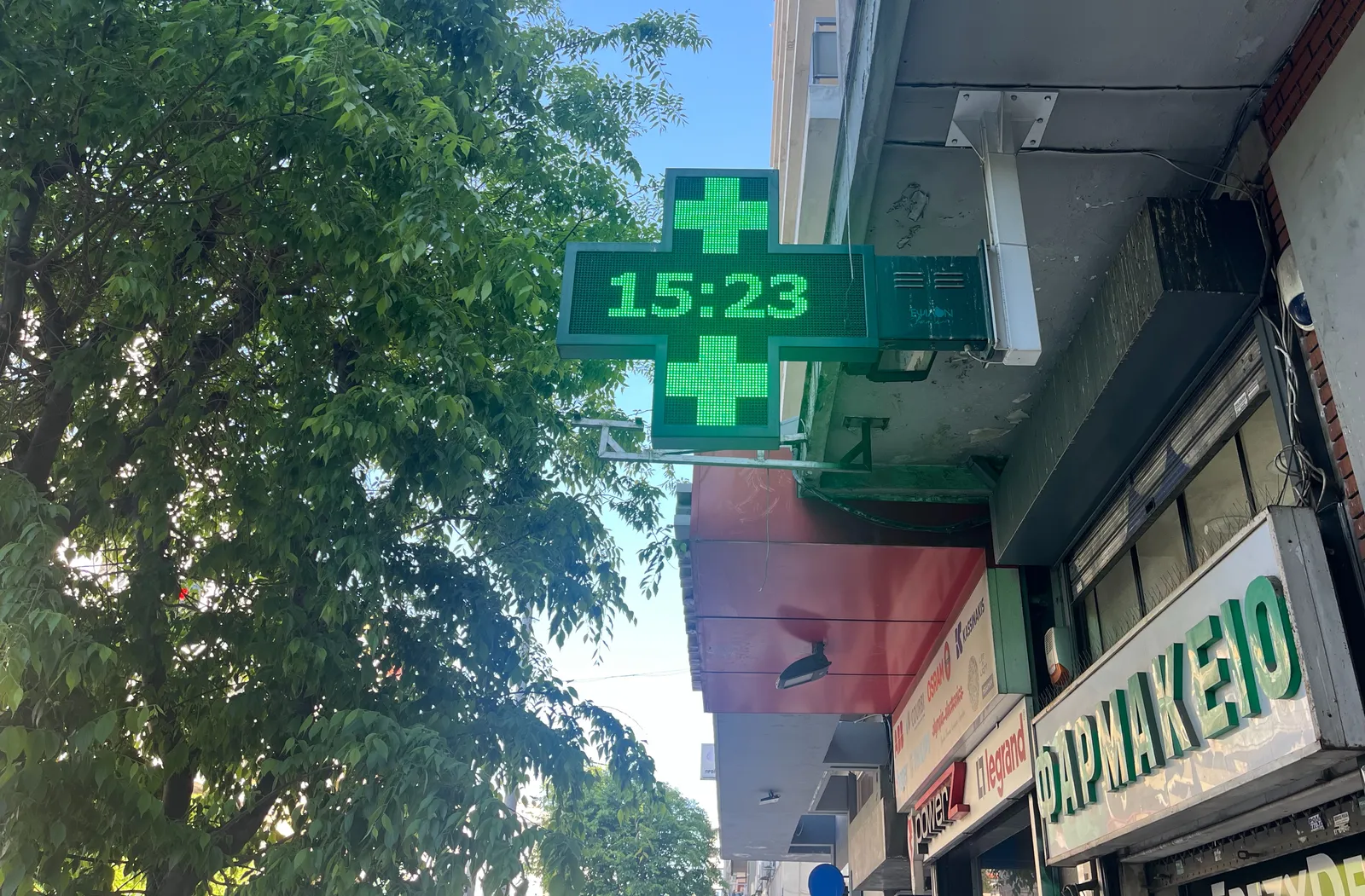 A large, green digital cross attached to the facade of a Greek pharmacy. On this cross the time it shown as 15:23