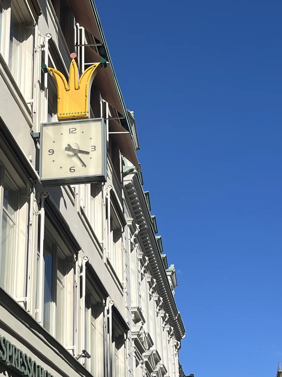 Attached to a neo-classicistish building there’s a clock with a cartoonish crown on top. The clock points at 24 minutes past three.