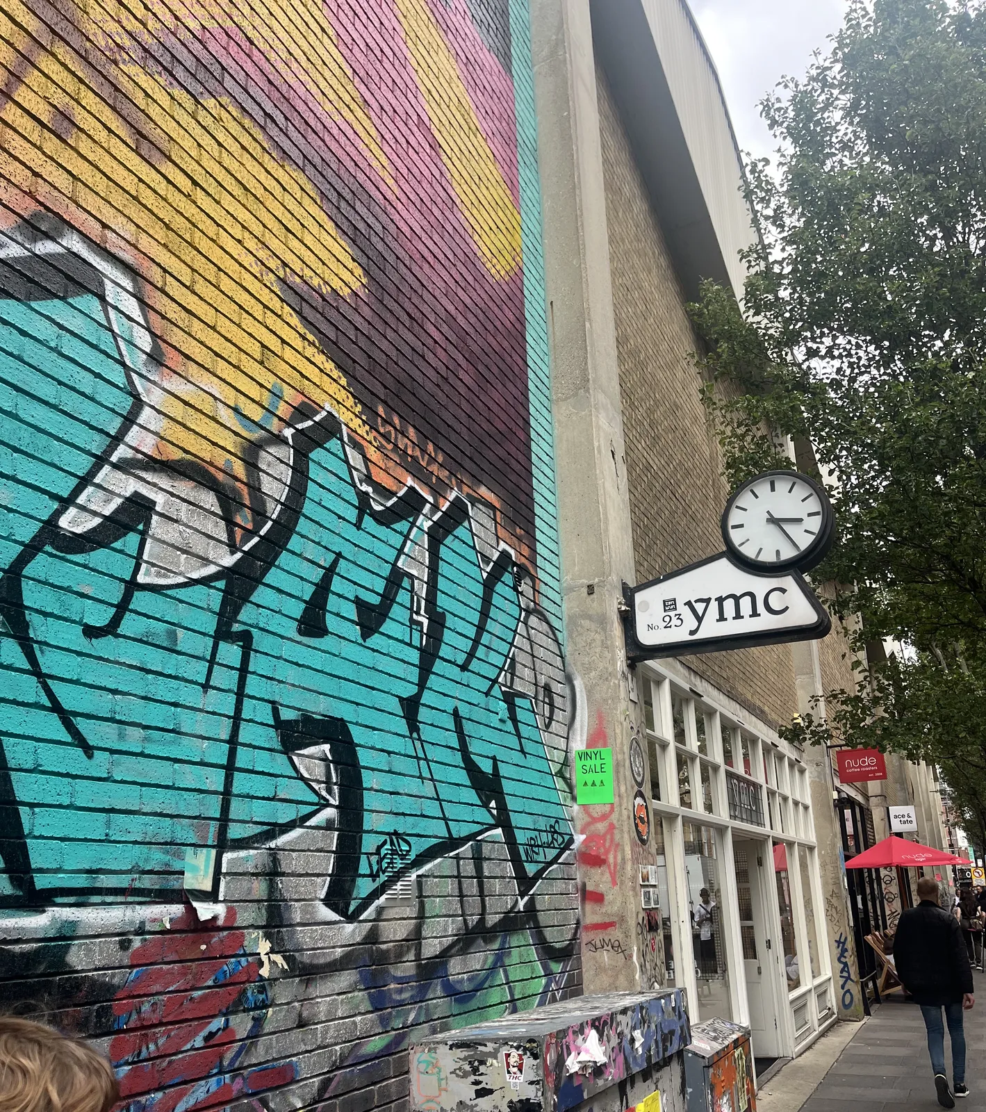 A high wall, filled with colourful graffiti. There’s a clock attached to this wall, above a sign that says No 23, YMC. The clock points at 25 minutes past three.