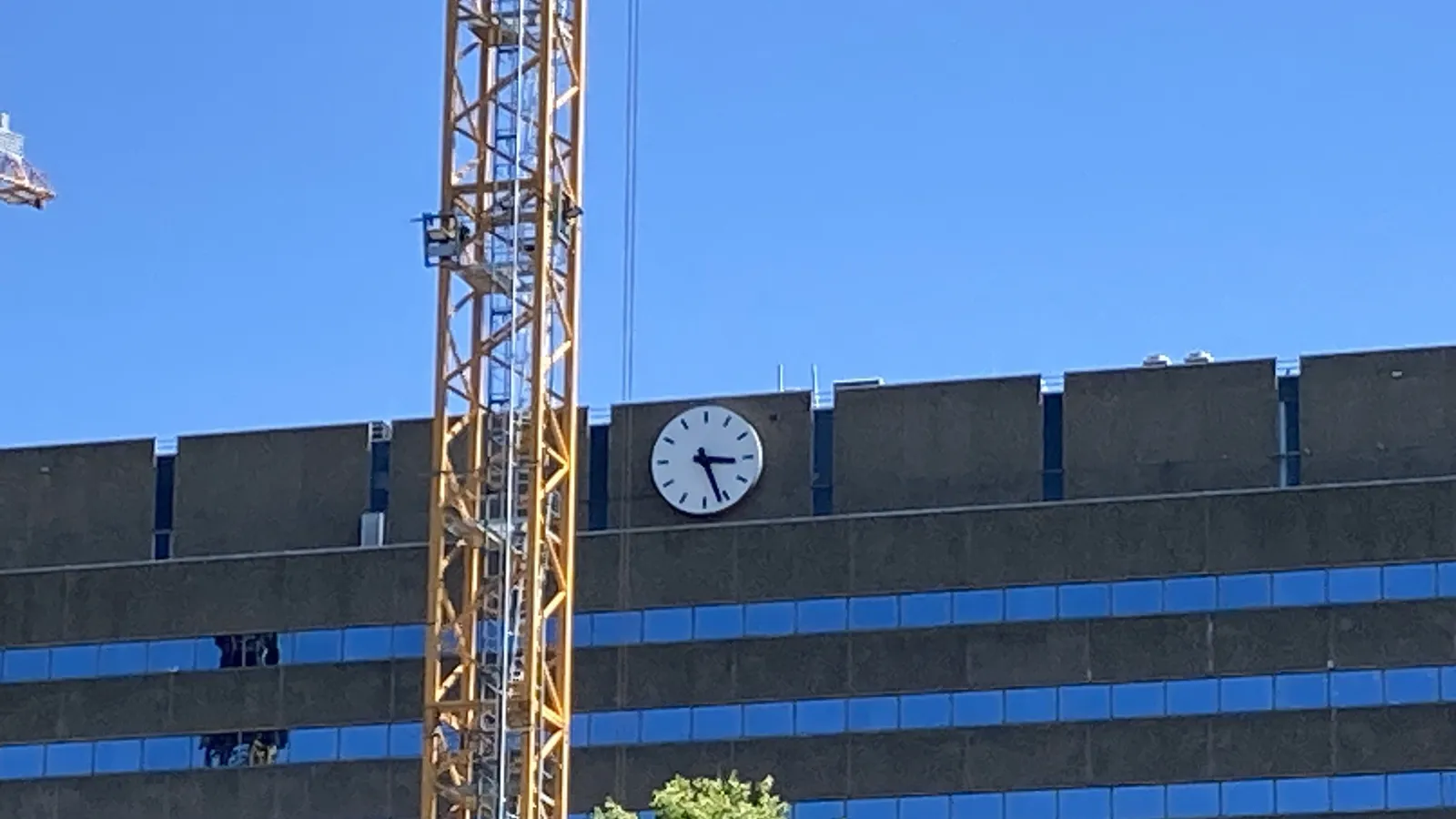 A yellow crane stands in front of a rather brutalist, concrete building with a clock on it. It points at 26 minutes past 3.