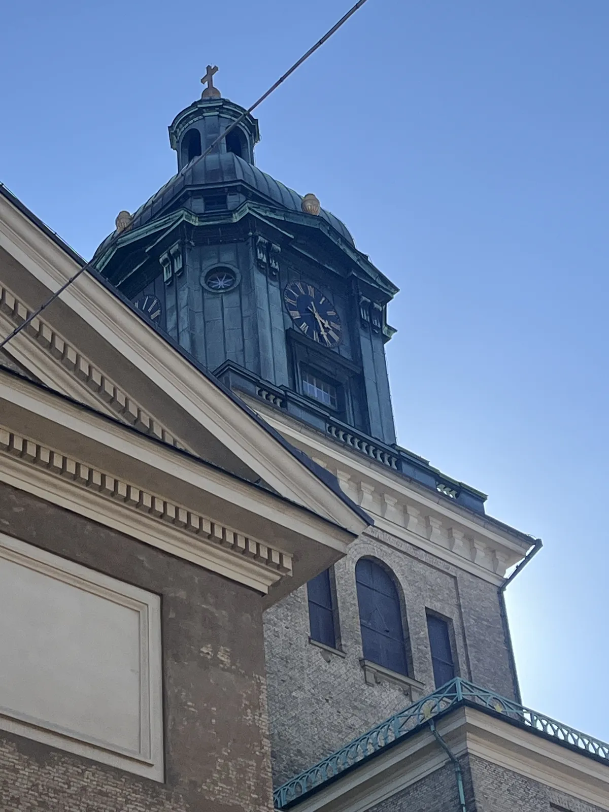 Looking up at the corner of a rather monumental, neoclassical, building with a brass dome. On top of the dome there’s a clock which points at 26 minutes past three.