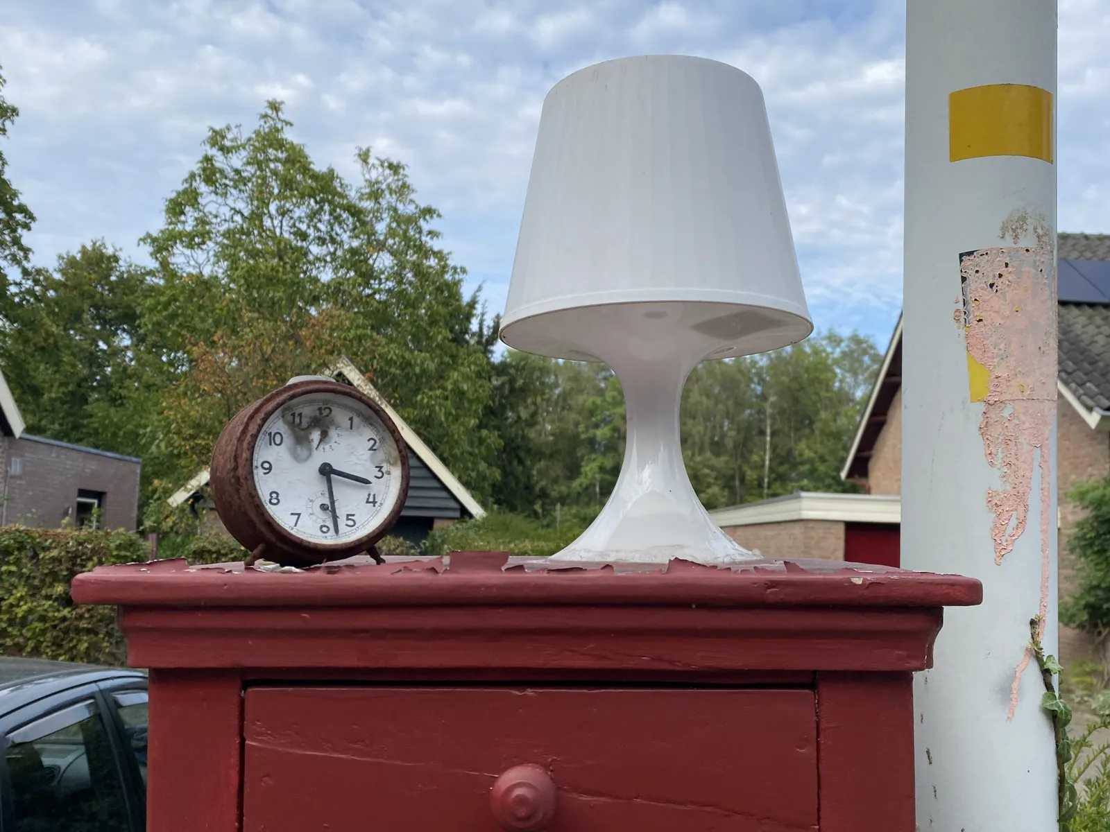 A small, red cupboard, standing outside, next to a pole. On this cupboard there’s a small, white plastic lamp, and an old, rusty alarm clock. It points at 28 minutes past 3.