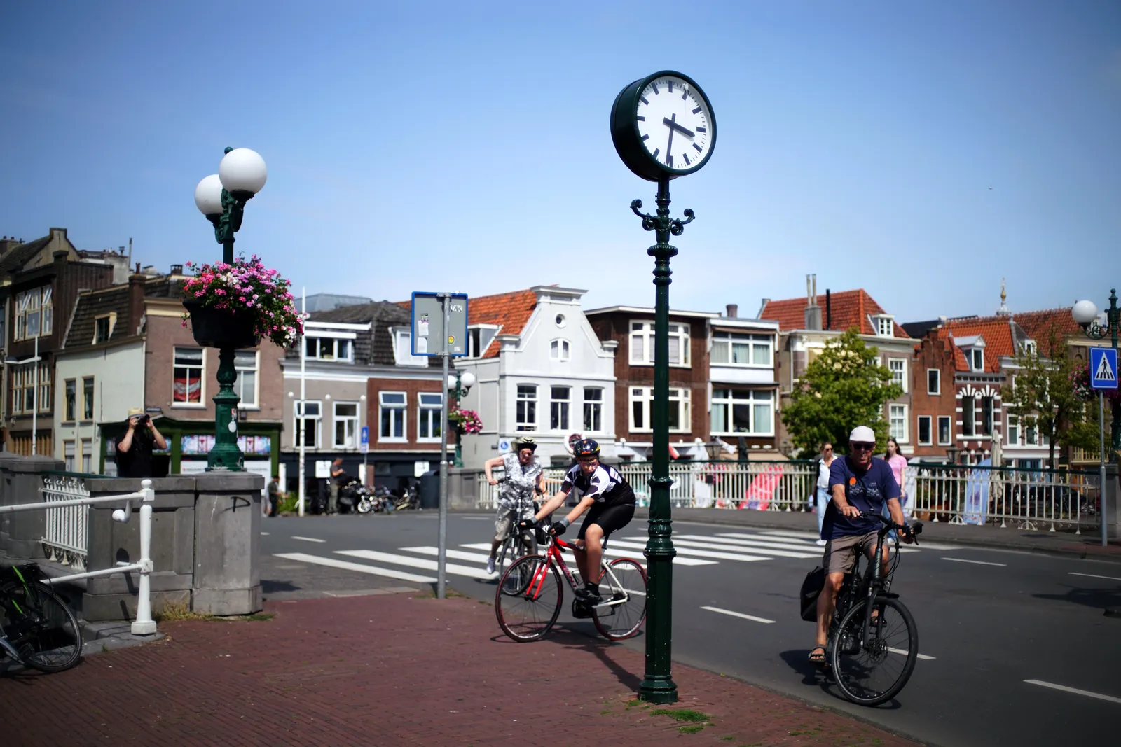 People on bikes going in all kinds of weird directions around a clock on a pole. Rather chaotic. Someone can be seen taking a picture of the clock, unaware of the chaos. The clock points at 31 past 3.