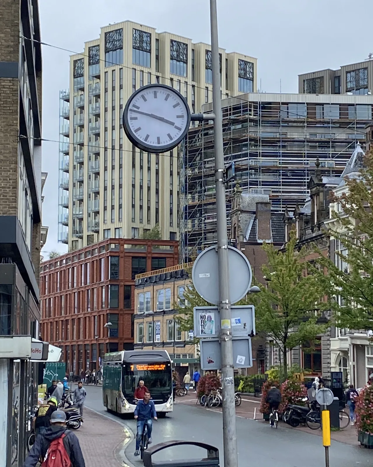 A clock on a pole in a cityscape. It points at 12 minutes to 4.