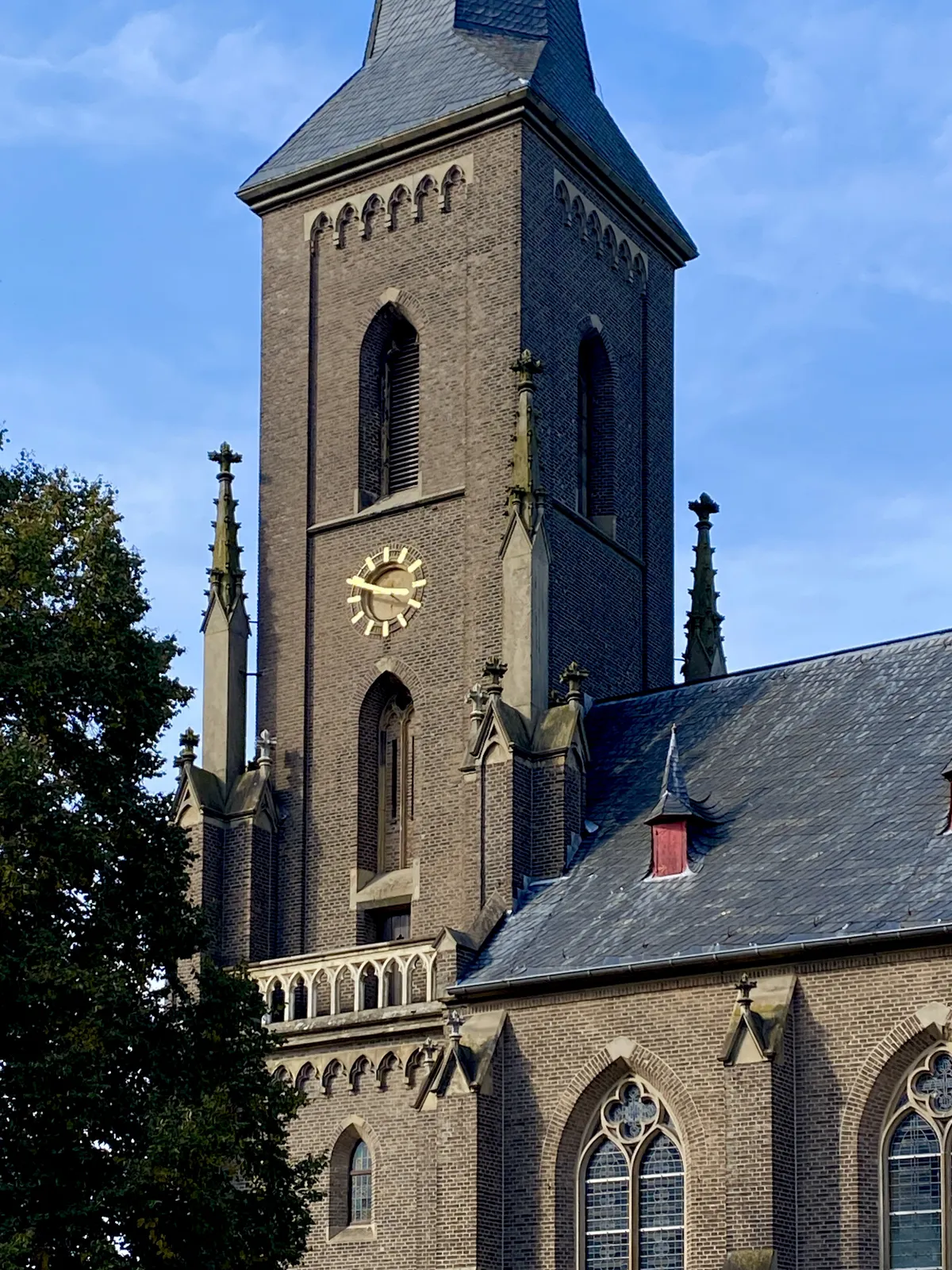 A brick church, with just a few, rather stern ornaments. It has a slate roof. The clock on its tower points at 11 minutes to 4.