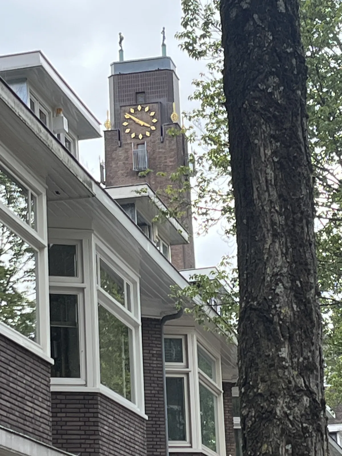 A tree trunk in the foreground, houses on the second plane, and a eye catching tower, with golden ornaments, and two statues of skinny, cheering figures on top in the background. There’s a clock on this tower, which points at 9 minutes to four.