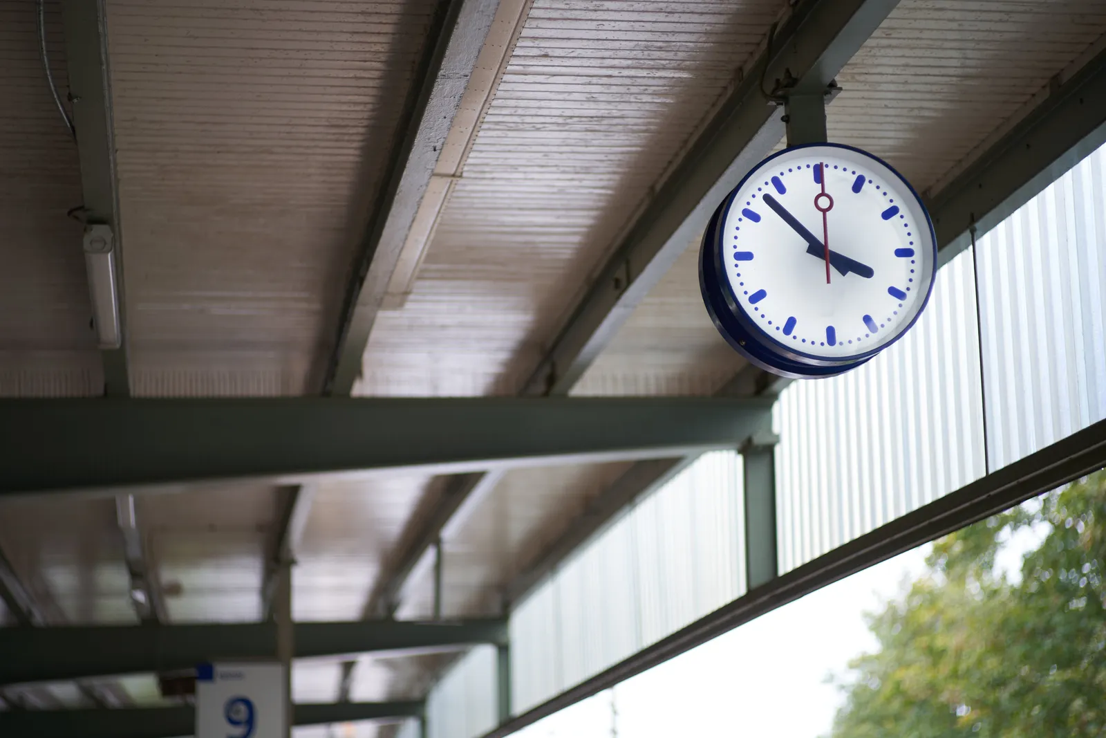 A clock, hanging from the ceiling in a train station. It points at 7 minutes to 4.