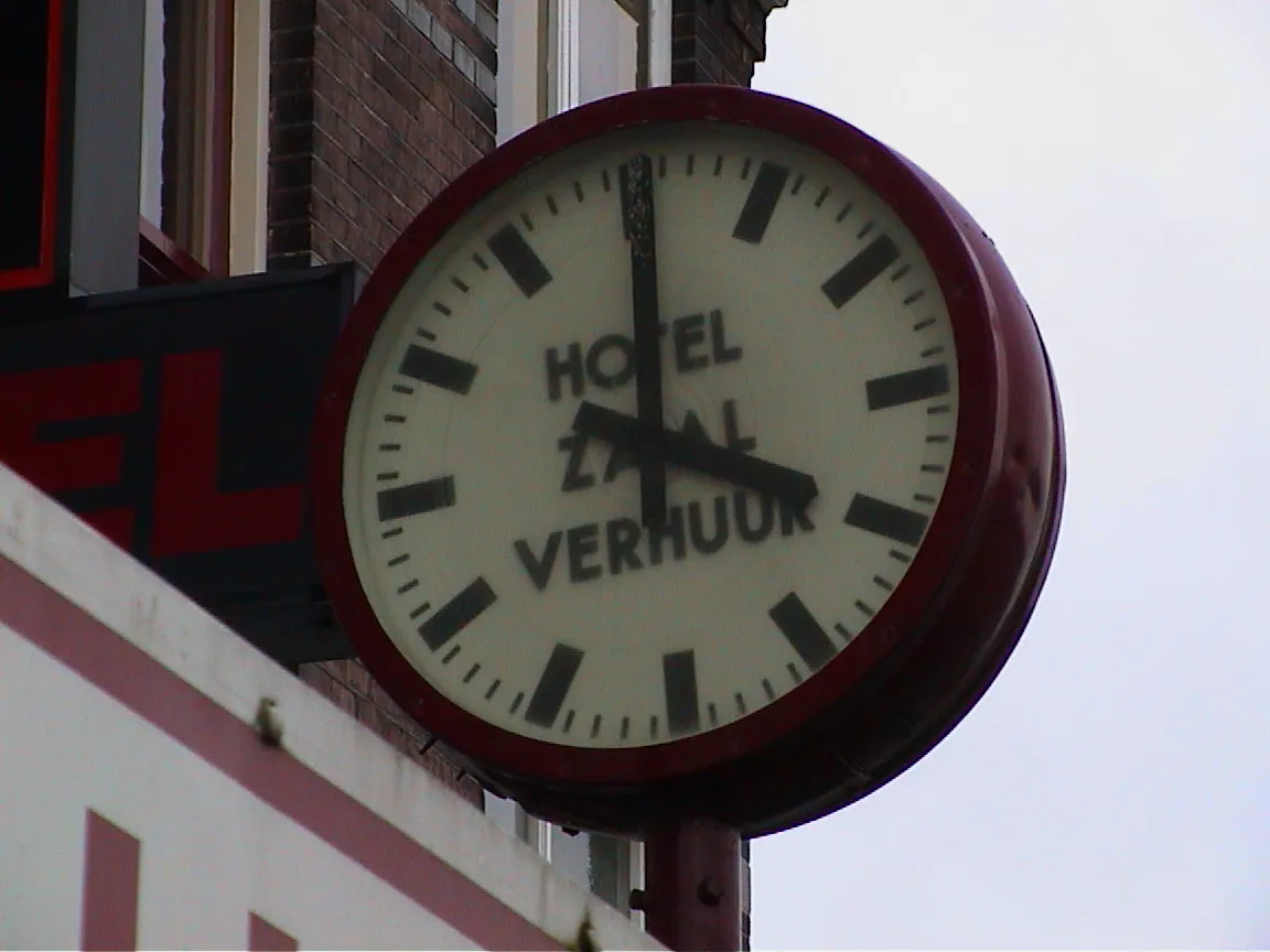 A clock attached to the wall. On its face it says Hotel Zaalverhuur, which means that it’s a hotel, and halls can be rented. It points at four o’clock.