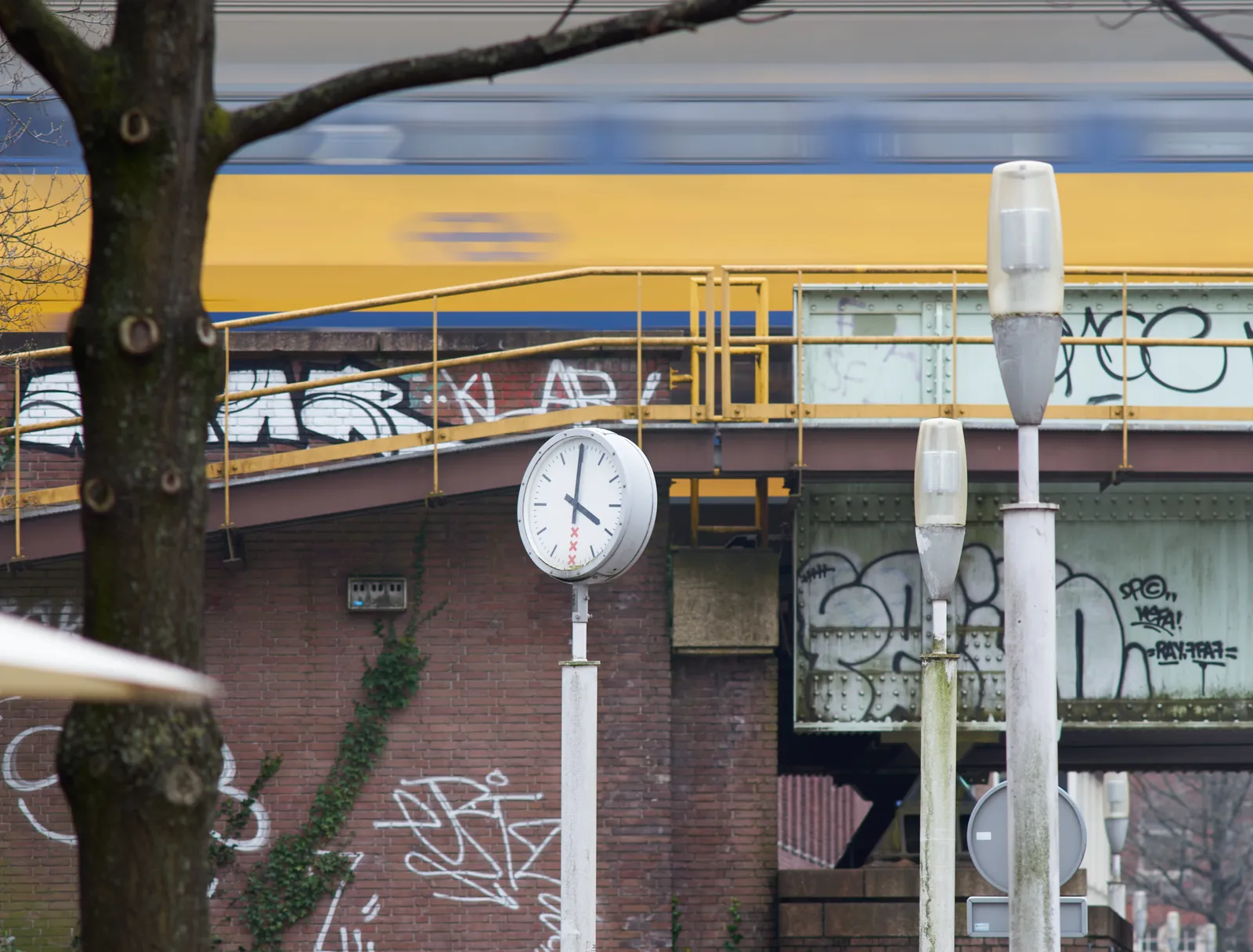 A train rolls over a bridge over a street. The clock in front of this bridge points at four o’clock.